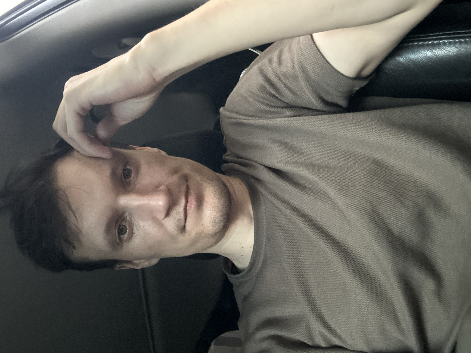 A medium shot shows a man sitting in a car, his head tilted slightly and his right hand resting on his forehead. He is wearing a brown, short-sleeved polo shirt. The background is dark, suggesting the interior of the vehicle. The man has short, dark hair and appears to be looking directly at the camera. The lighting is dim, casting soft shadows. There is no visible indication of the city of Kamianets-Podilskyi, Ukraine.