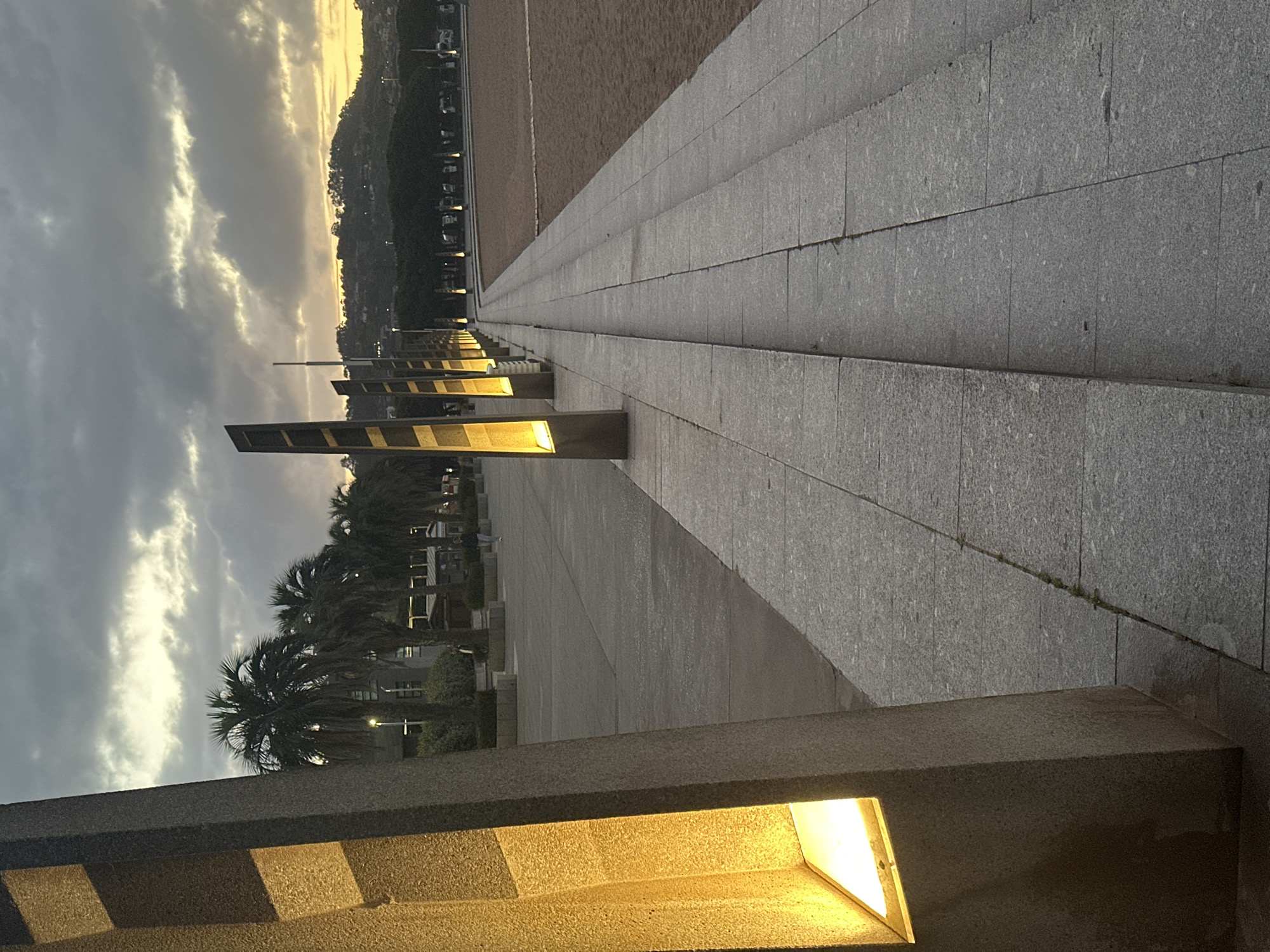 A wide, grey stone promenade, stretching diagonally from the bottom-left to the middle-right of the frame, dominates the scene. The promenade is composed of large, rectangular stone slabs with visible seams. Along its right edge, a series of modern, rectangular light fixtures are integrated into a low stone wall or structure, casting a warm, bright yellow glow onto the path and creating strong linear shadows.
To the left of the promenade, a vast, flat expanse of sandy ground extends into the distance, possibly indicating a beach or a large open area. In the mid-ground, further to the left, several dark green palm trees are visible, silhouetted against the sky.
The background on the far right features a distant hill or mountain, covered in vegetation and dotted with scattered buildings, suggesting a coastal town or city. The sky above is partially cloudy, with lighter, warm-toned patches breaking through darker clouds, characteristic of either dawn or dusk. The artificial lights are prominently lit, reinforcing the low-light conditions. The overall mood is calm and urban, with no people or specific activities depicted. No text is visible in the image. The setting is a paved waterfront area in Gijón, Spain.