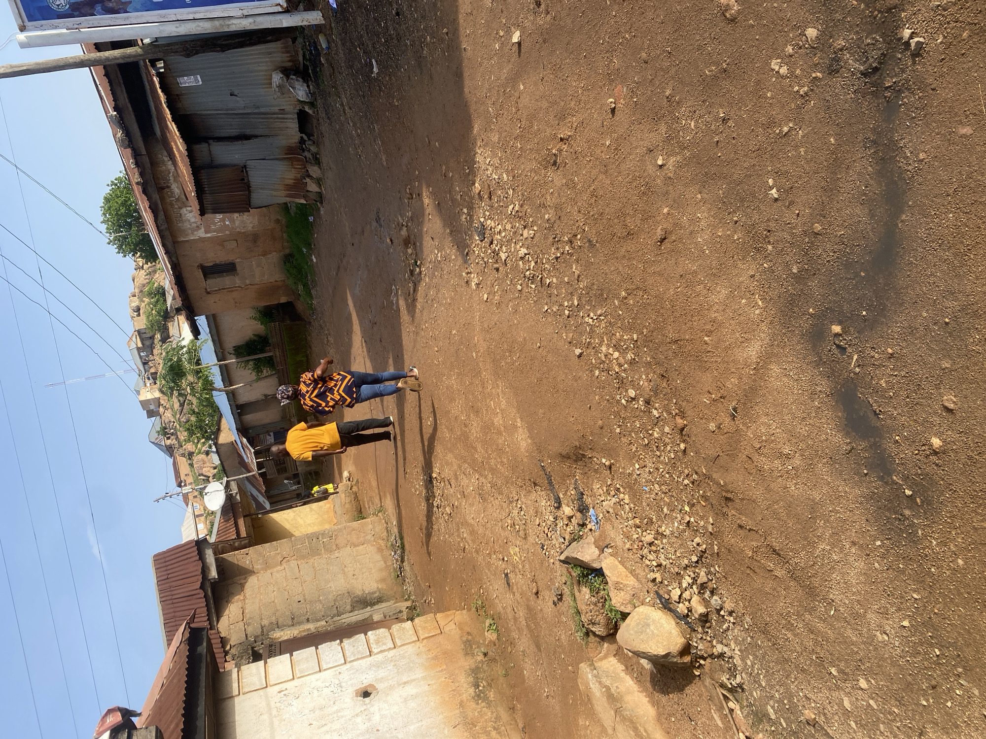 The image depicts a street scene in Jos, Nigeria. Two individuals are visible walking on a dirt path. One person wears a patterned shirt and blue jeans, while the other is dressed in a yellow t-shirt and dark pants. They are walking away from the camera, towards a cluster of buildings. The buildings appear to be made of brick and corrugated metal, suggesting a somewhat informal urban environment. Some of the structures show signs of wear and tear.
The ground is primarily composed of reddish-brown dirt, with scattered rocks and some patches of vegetation. There are visible tire tracks on the dirt path, indicating vehicular traffic. A shadow stretches across the ground, suggesting it is daytime, likely mid-morning or afternoon given the angle of the light. The sky is clear and blue, hinting at fair weather.
In the background, more buildings are visible, some situated on a rocky incline. Power lines are strung across the sky, a common sight in many urban areas. A satellite dish is mounted on one of the buildings. The overall impression is one of a typical day in a bustling, developing urban landscape. No text is clearly legible in the image.