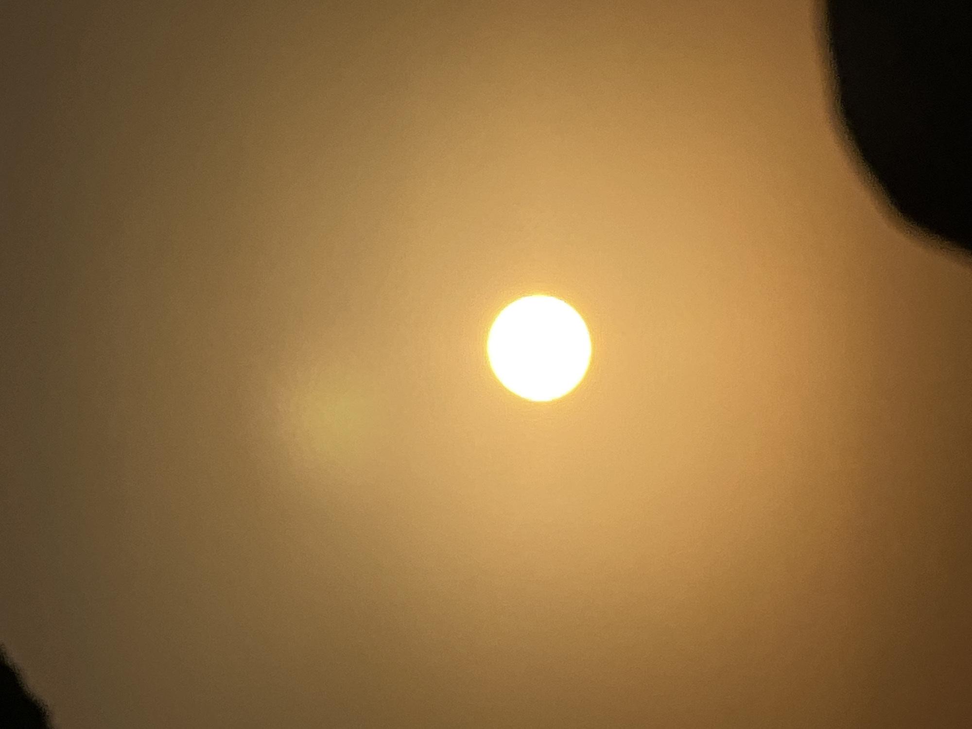 A bright, circular sun is centered in an amber-toned sky, likely during sunrise or sunset. The sky is hazy, obscuring any details of the horizon or surrounding environment. In the lower right corner, a blurred, dark silhouette, possibly a person's head or shoulder, is visible, suggesting the photograph was taken from a low vantage point. The overall scene depicts a natural phenomenon, with the sun as the dominant subject, set against a diffused, atmospheric background. The location in Kuta, Indonesia is not discernible from the visual information presented.