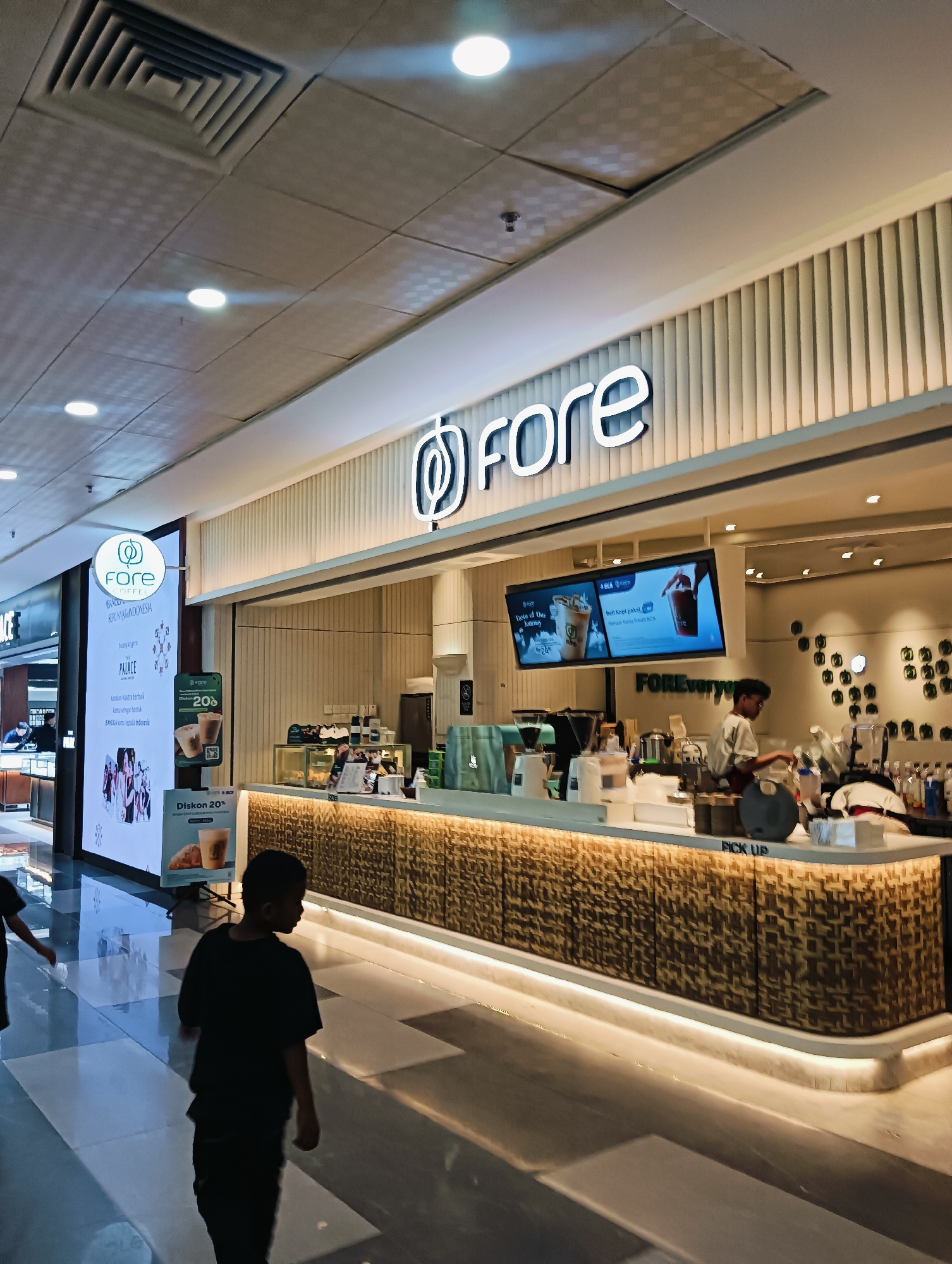 The image depicts the interior of a modern coffee shop, likely "Fore Coffee," located in Batam, Indonesia. The shop features a clean, minimalist design with a prominent white counter adorned with a textured, patterned facade. Behind the counter, staff members are visible preparing drinks, with various coffee machines and equipment on display. Digital screens above the counter showcase menu items and promotions. A circular illuminated sign with the "Fore Coffee" logo is visible on the left side of the frame. In the foreground, two young boys, one silhouetted and walking away from the camera, are present. The lighting suggests it's daytime, and the polished floor reflects the overhead lights and shop interior. The overall ambiance is that of a bustling, contemporary retail establishment.