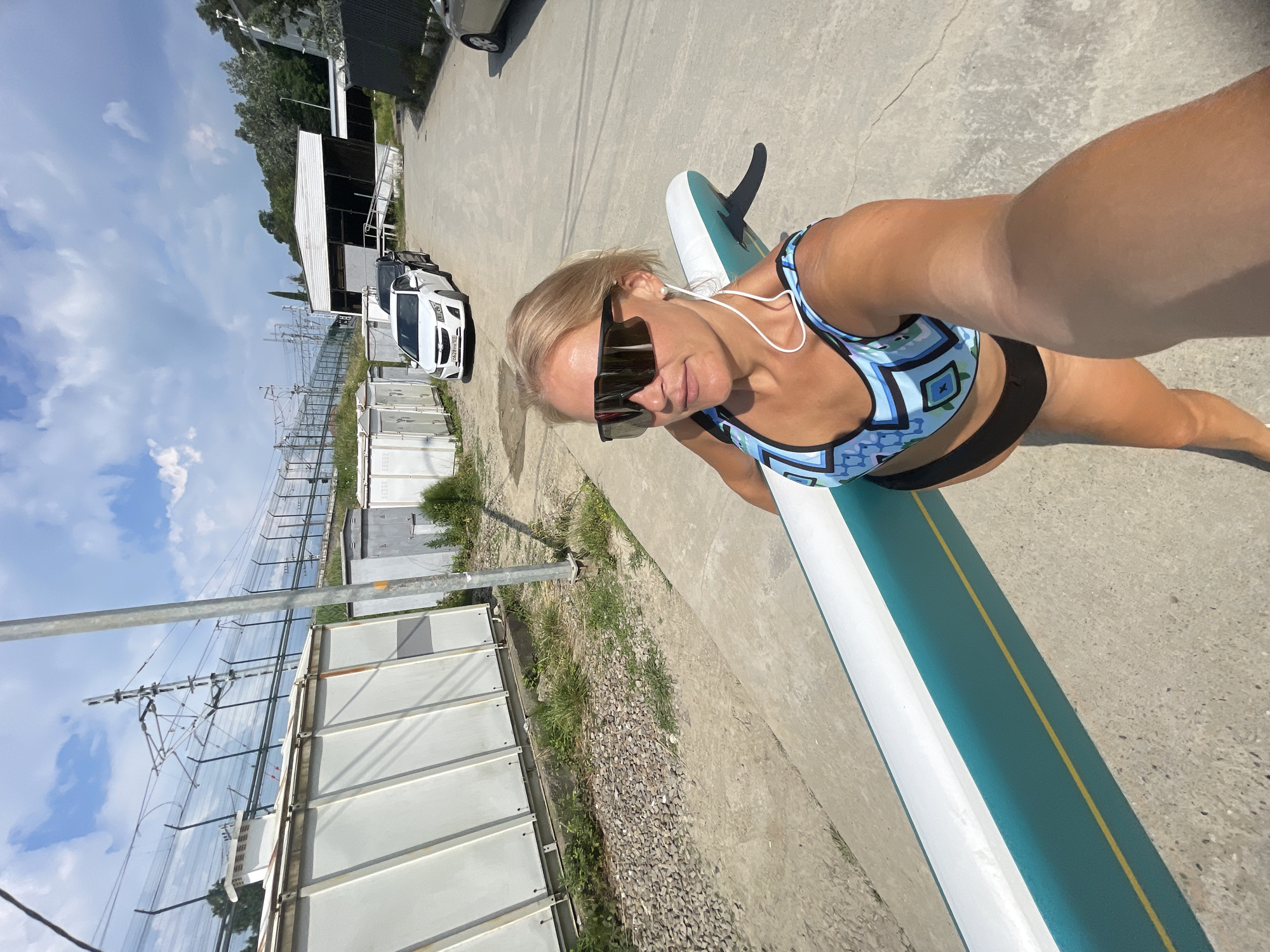 The media file shows a woman standing on a paddleboard on a sunny day. She is wearing a blue and white patterned bikini top and black bottoms. She has on large, dark sunglasses and white earbuds. The paddleboard is white with a teal stripe and a yellow line down the center. She appears to be posing for a selfie, as the camera is pointed towards her.
The scene is an outdoor, industrial-looking area. In the background, there are several white shipping containers and a white car. There are also power lines and poles, suggesting proximity to an electrical substation or similar infrastructure. The ground is paved with concrete. Lush green trees and grass are visible in some areas, contrasting with the concrete and industrial elements. The sky is blue with scattered white clouds, indicating fair weather and daylight. The overall impression is that of a casual outing in a somewhat unconventional location.
The woman's pose and smile suggest a positive and confident demeanor. The image is taken from a slightly elevated angle, emphasizing the paddleboard and the woman. The orientation of the image is rotated, with the woman and paddleboard appearing to be on the left side and running diagonally towards the right.
No visible text is present in the media file. The location context of Sochi, Russia, is provided externally and not discernible from the image itself.