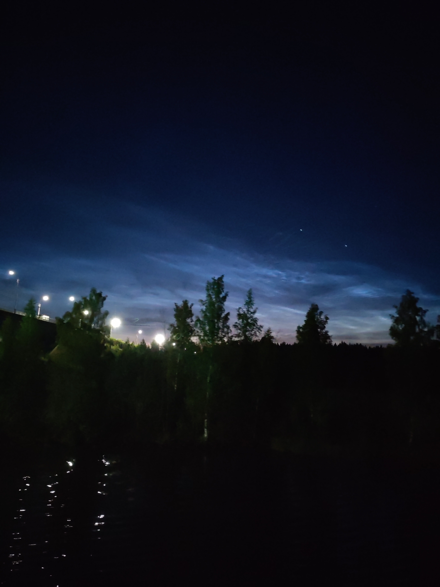 A nighttime photograph captures a dark, silhouetted landscape with a deep blue sky above. The sky features wispy, luminous clouds, possibly noctilucent clouds, illuminated by an unseen light source. Two faint celestial bodies are visible in the upper right portion of the sky. In the mid-ground, a line of trees forms a dark silhouette against the brighter sky. Several streetlights are visible, some appearing as bright white circles, interspersed among the trees, suggesting the presence of a road or pathway. The foreground is dominated by a dark, reflective body of water, likely a river or lake, with streaks of light from the streetlights shimmering on its surface. The overall scene suggests a rural or semi-urban environment at twilight or night, with artificial light sources interacting with natural atmospheric phenomena. The location is identified as Krapivino, Russia. There are no people or discernible actions depicted in the image.