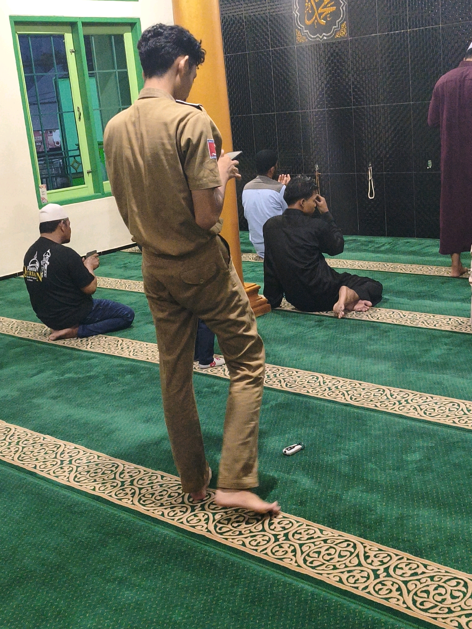 The image depicts the interior of a mosque in Samarinda, Indonesia, featuring green carpeted floors with decorative gold patterns. Several men are present, most of whom appear to be engaged in prayer or seated on the carpet. In the foreground, a young man in a light brown uniform stands, holding a mobile phone. To his left, another man wearing a white skullcap and a black t-shirt with text on the back is seated. Further into the mosque, other individuals are visible, including one in a blue shirt and another in a black garment, both seated on the floor. The background shows a black tiled wall with decorative Islamic calligraphy above. A man in a dark maroon robe stands at the far right of the frame. A green-framed window is visible on the left side of the image. The overall impression is of a communal religious gathering.