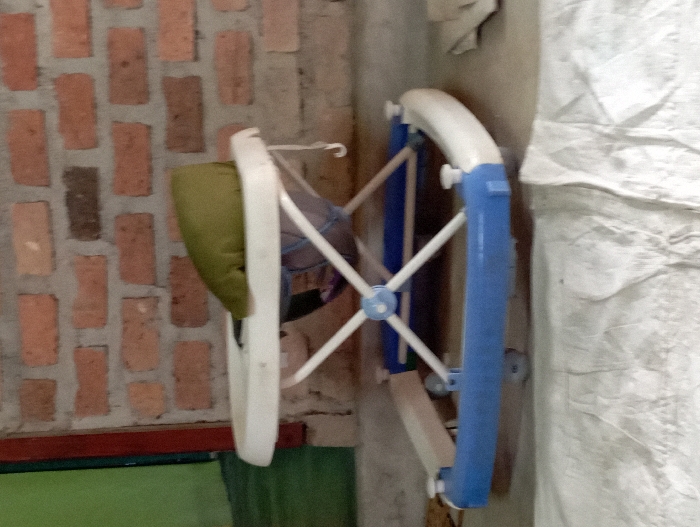 The image, taken in Pubian, Indonesia, depicts a baby walker, likely folded or collapsed, propped vertically against a wall. The walker consists of a white plastic frame with prominent blue plastic components on its right side, forming a curved handle or support. A green fabric cushion or seat is partially visible on the left side of the walker's frame.

The background features a rustic brick wall on the left, composed of reddish-brown bricks with visible mortar lines. To the right of the walker, a large, textured white fabric object, possibly a storage bag or draped material, leans against another wall or surface. The lower left section of the image shows a reddish-brown horizontal strip, potentially a wooden beam or baseboard, above a muted green area, which might be flooring or a lower part of a wall. The lighting suggests an indoor setting, with no indication of natural light or time of day. No individuals are present, and no particular activity is taking place beyond the storage of the item. There is no visible text in the image.