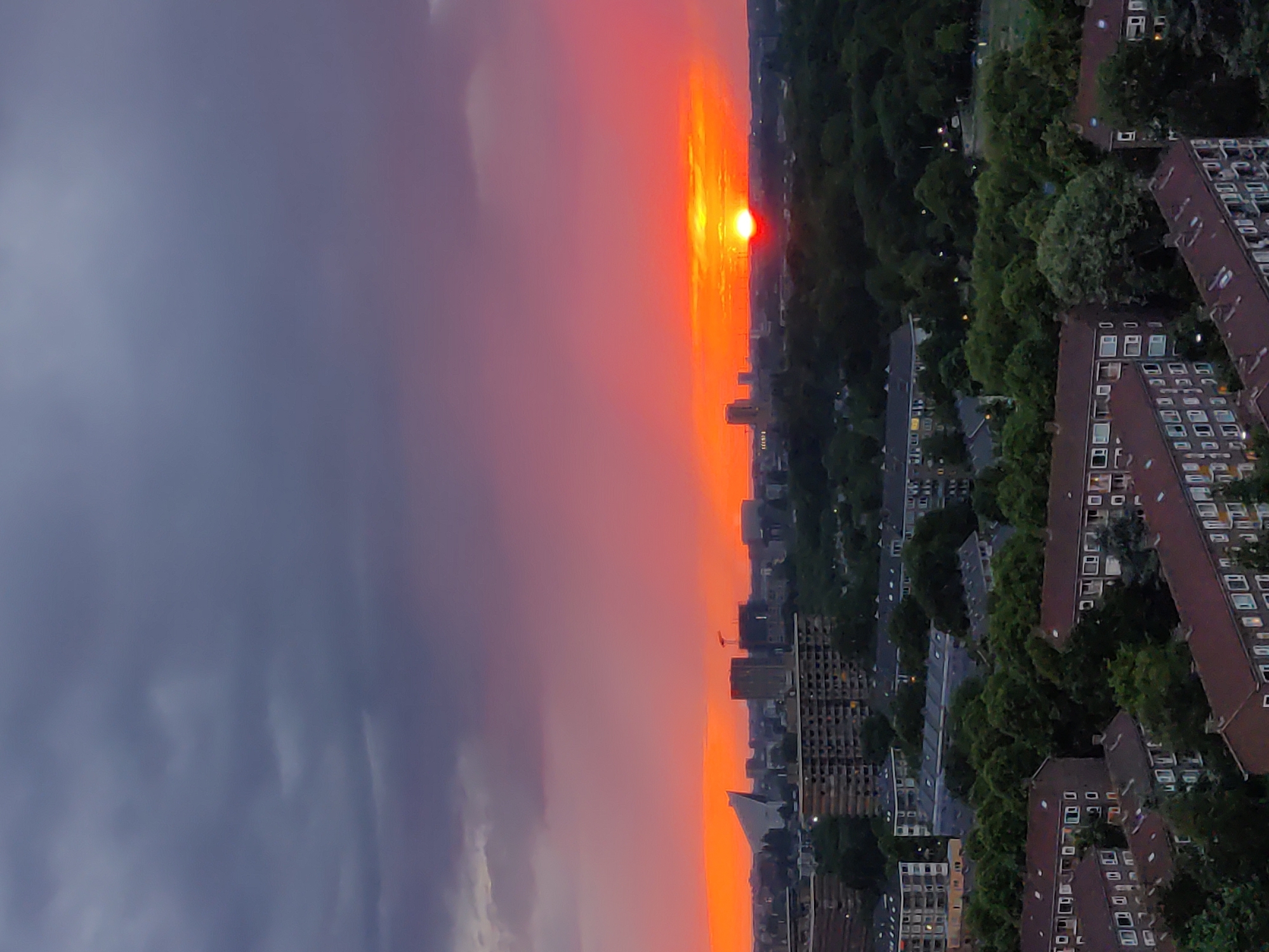This image captures a vibrant sunset over the city of Rijswijk, Netherlands. The sky is a dramatic canvas of deep grays and purples at the top, transitioning to a brilliant gradient of pinks and fiery oranges near the horizon. The sun, a bright orange orb, is prominently visible, just beginning to dip below the skyline.
The cityscape below is a mixture of residential buildings and green spaces. In the foreground, several multi-story apartment buildings with reddish-brown roofs are visible. These buildings feature numerous windows and balconies. Further in the background, more apartment blocks and some taller structures, possibly office buildings or hotels, punctuate the landscape. A distinctive, modern white building with a triangular roof is also visible on the left side of the city. The horizon is lined with trees, forming a dense, dark green band that separates the sky from the urban environment. A construction crane stands tall among the buildings, suggesting ongoing development in the city. The overall scene is one of an urban twilight, bathed in the warm, dramatic light of the setting sun. No people are visible in the image.