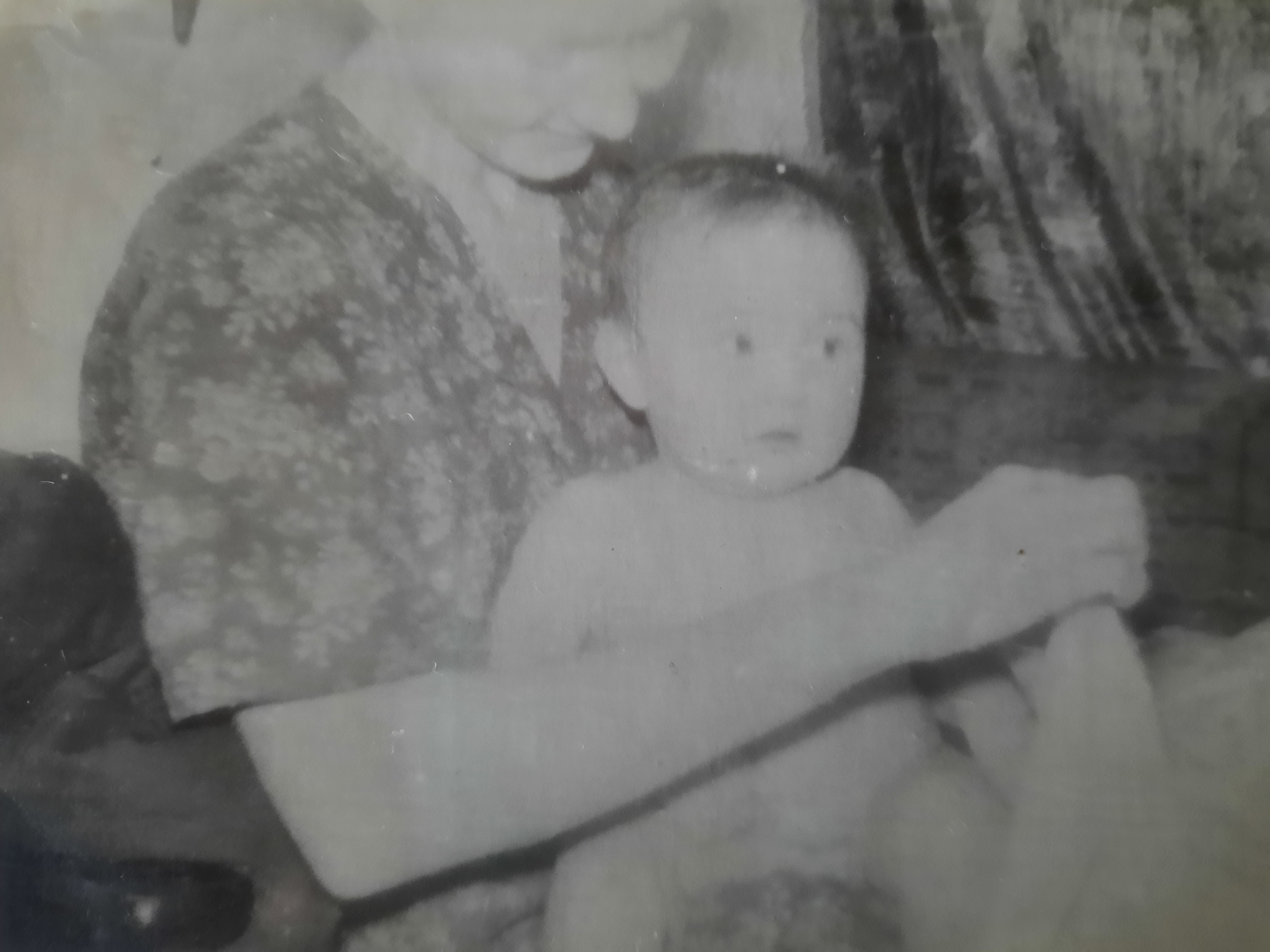 The image appears to be a black and white photograph of a baby or young child being held by an older individual, presumably a woman. The woman is wearing a dress or top with a floral pattern. The baby is positioned in front of her, and one of the woman's arms is visible, holding the baby. Another individual is interacting with the baby's hand. The location is unknown as the image only shows the people and objects in the frame. The environment behind them is blurry and indistinct. The image's quality is low, with significant fading and contrast issues, common in old photos.