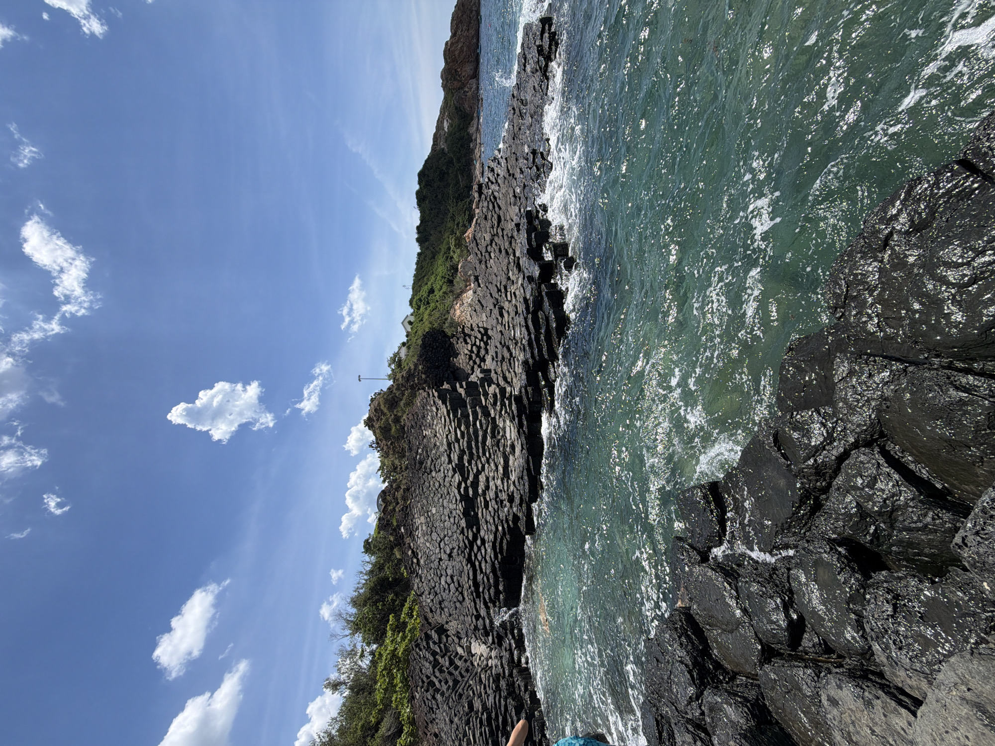 The image shows a scenic coastal landscape in Phú Lương, Vietnam. In the foreground, dark, hexagonal basalt rock formations emerge from the clear, turquoise ocean water. The water is slightly choppy, with white foam breaking around the rocks.
Behind the foreground rocks, a larger formation of similar hexagonal basalt columns rises up the hillside. These columns are stacked and layered, creating a textured, stepped appearance. Green vegetation covers the slopes of the hill, with some trees and bushes visible. A white building is situated on top of the hill in the distance.
The sky above is a vibrant blue, with scattered white clouds. The lighting suggests it is daytime, with the sun casting highlights on the wet rocks and the surface of the water. A person's arm, wearing a blue and white patterned garment, is visible on the far left of the image, reaching towards the water. The overall scene depicts a natural geological formation meeting the sea under a bright sky.