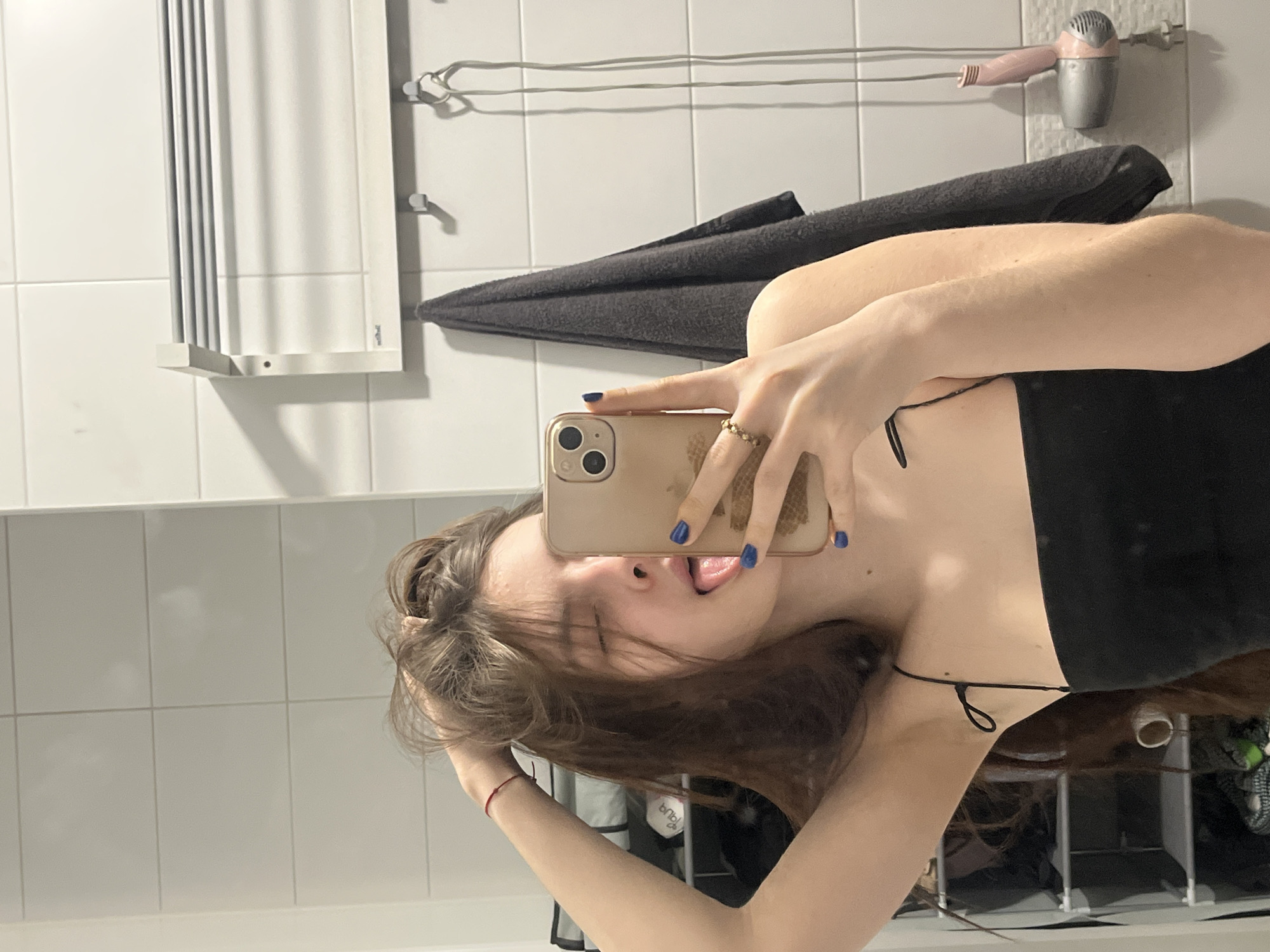 A young woman with long brown hair is taking a selfie in a bathroom. She is wearing a black top with thin straps. Her tongue is slightly out. Her fingernails are painted blue. She is holding a gold-colored iPhone with three lenses. Behind her are white tiled walls and a towel rack with a gray towel and a pink and silver hairdryer. There is also a white towel warmer. The location is most likely inside a residence, but there is no indication it is in Saint Petersburg, Russia.