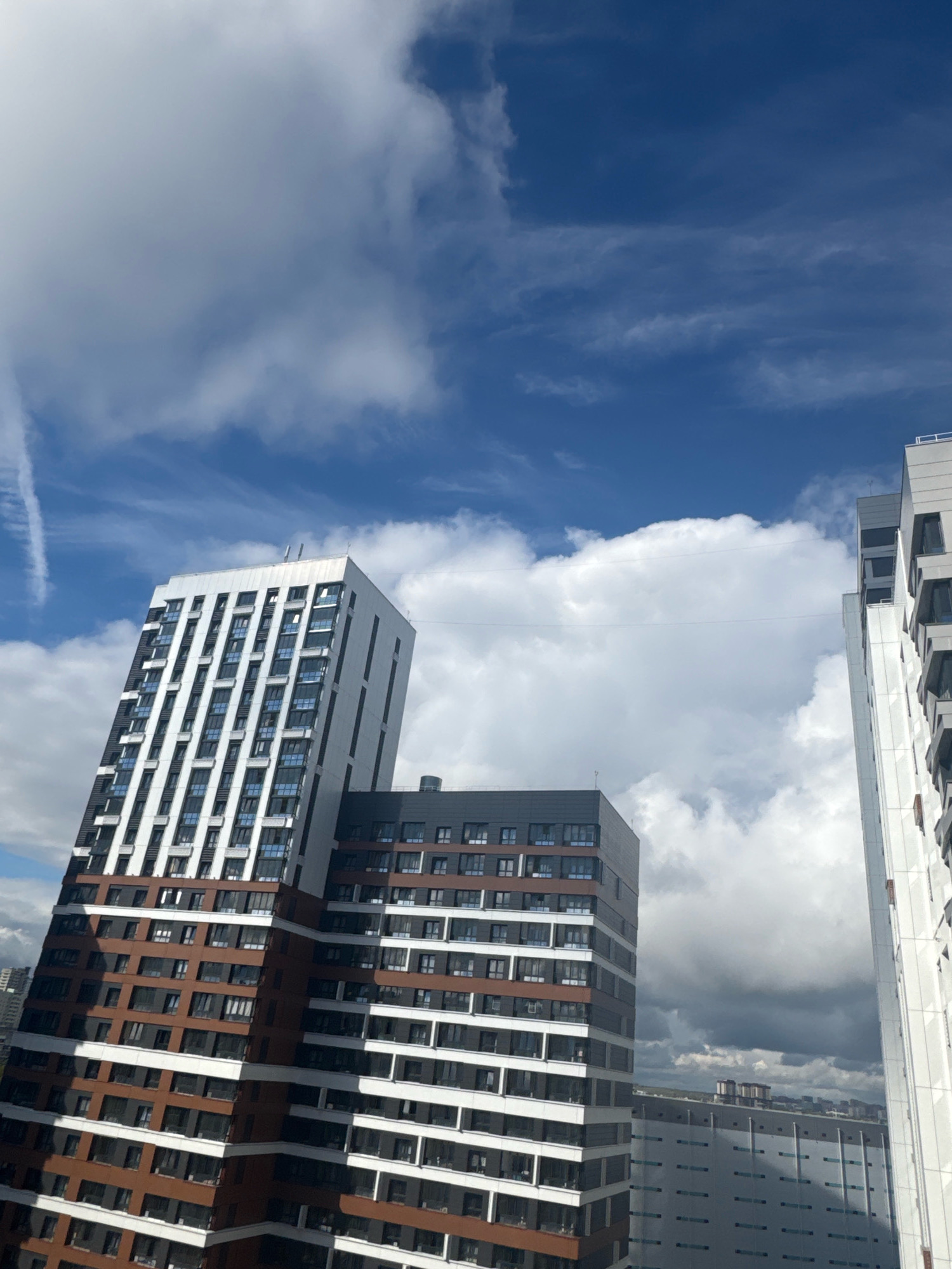 An urban vista features modern high-rise buildings under a partly cloudy sky. The buildings are characterized by their contemporary architectural style, with repeating window patterns and contrasting facade materials, including dark brown and white sections. The sky is a deep blue, punctuated by large, fluffy white clouds and some thinner, wispy clouds. In the distance, other buildings and structures are visible, suggesting a developed urban environment. The perspective is from an elevated viewpoint looking upwards, emphasizing the scale of the buildings against the sky. There are no people or animals visible in the frame. The overall scene captures a typical day in a city with a focus on its architectural landscape and the sky. It is not possible to determine the specific location in Novosibirsk, Russia from the provided image.