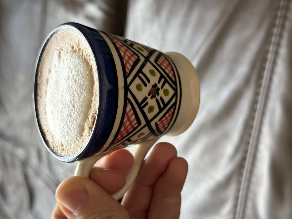 A light-skinned right hand is gripping the handle of a small, decorative ceramic cup. The cup features a dark blue rim and a white base, with its body adorned in a geometric pattern of red cross-hatching, dark blue outlines, and green and black accent dots on a white background. The cup contains a light brown liquid topped with a thick layer of white, frothy foam. The background is a softly blurred, light gray or beige upholstered surface with visible horizontal stitching lines, suggesting a couch or similar furnishing. The scene is located in Sofia, Bulgaria.