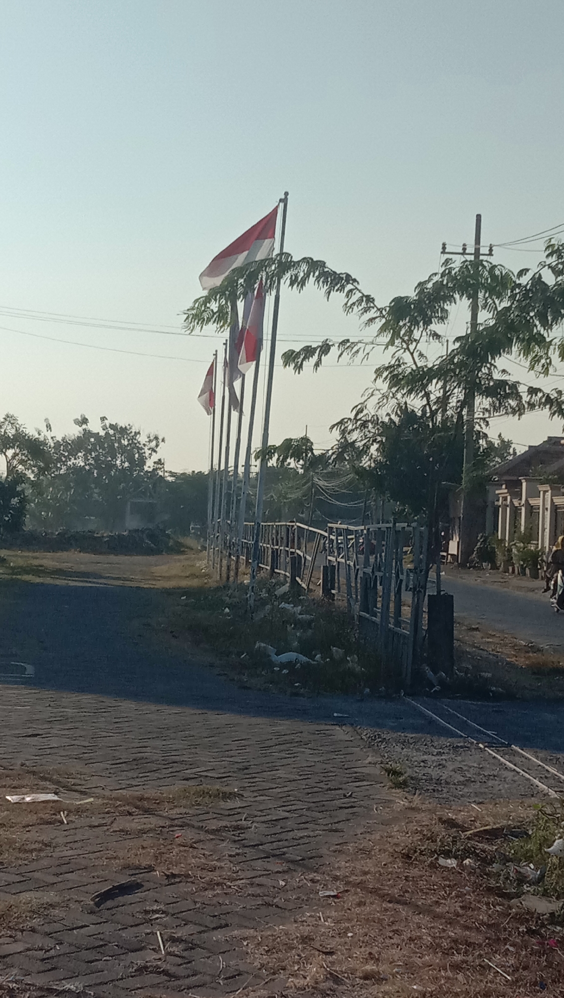 The image depicts a road in Curah Dukuh, Indonesia, on a bright, clear day. In the foreground, a paved surface with interlocking brick patterns transitions to a darker asphalt road. To the right, a low wooden fence runs along the edge of the road, bordered by sparse vegetation and scattered debris. A row of flagpoles with Indonesian flags, displaying red and white horizontal stripes, stands prominently along the fence line, some partially obscured by the leaves of overhead trees. Beyond the flags, a two-story building with a tiled roof and white pillars is visible on the right side of the road, suggesting a residential or commercial area. The background reveals more trees and a hazy sky, with power lines crossing the upper portion of the frame. The scene appears quiet, with no individuals clearly visible, though the presence of parked motorcycles to the right hints at activity. The overall impression is one of a typical roadside scene in a rural or semi-urban Indonesian setting.