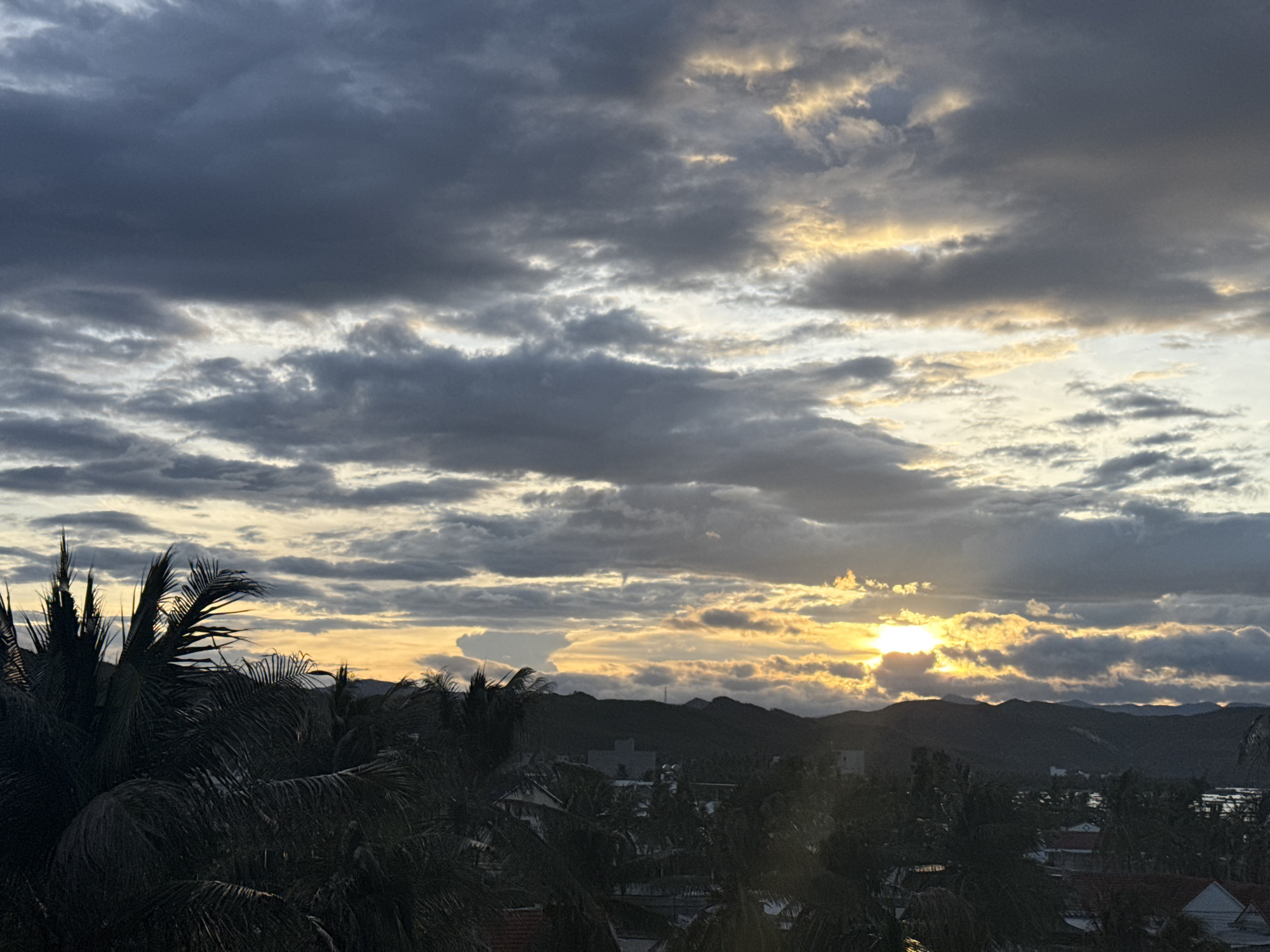 An overcast sunset casts a golden glow through dramatic clouds over a silhouetted landscape of palm trees, mountains, and buildings. The sun's rays pierce through the thick, grey clouds, creating a dramatic effect. The foreground is dominated by the dark, lush fronds of palm trees, obscuring much of the settlement below. In the mid-ground, dark, hilly terrain is dotted with buildings, some with red roofs. The sky is a complex tapestry of dark grey and lighter grey clouds, with bright yellow and orange hues where the sun is breaking through.