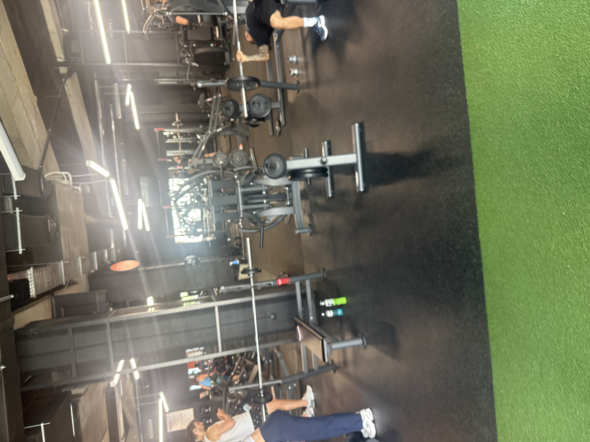 The image depicts an indoor gym setting. It appears to be in a commercial gym facility, with a variety of weightlifting equipment, including squat racks, barbell benches, and Smith machines. There are at least two individuals present. One person is performing a barbell exercise in a squat rack, while another is lying on a bench, presumably weightlifting. The gym has a black floor and green artificial turf in one section. The lighting is bright, with fluorescent lights mounted on the ceiling. The overall setting appears to be a standard, functional weightlifting area within a gym. The location is not discernible from the image itself, so it is not possible to confirm if it is in Plovdiv, Bulgaria, or any other specific city.