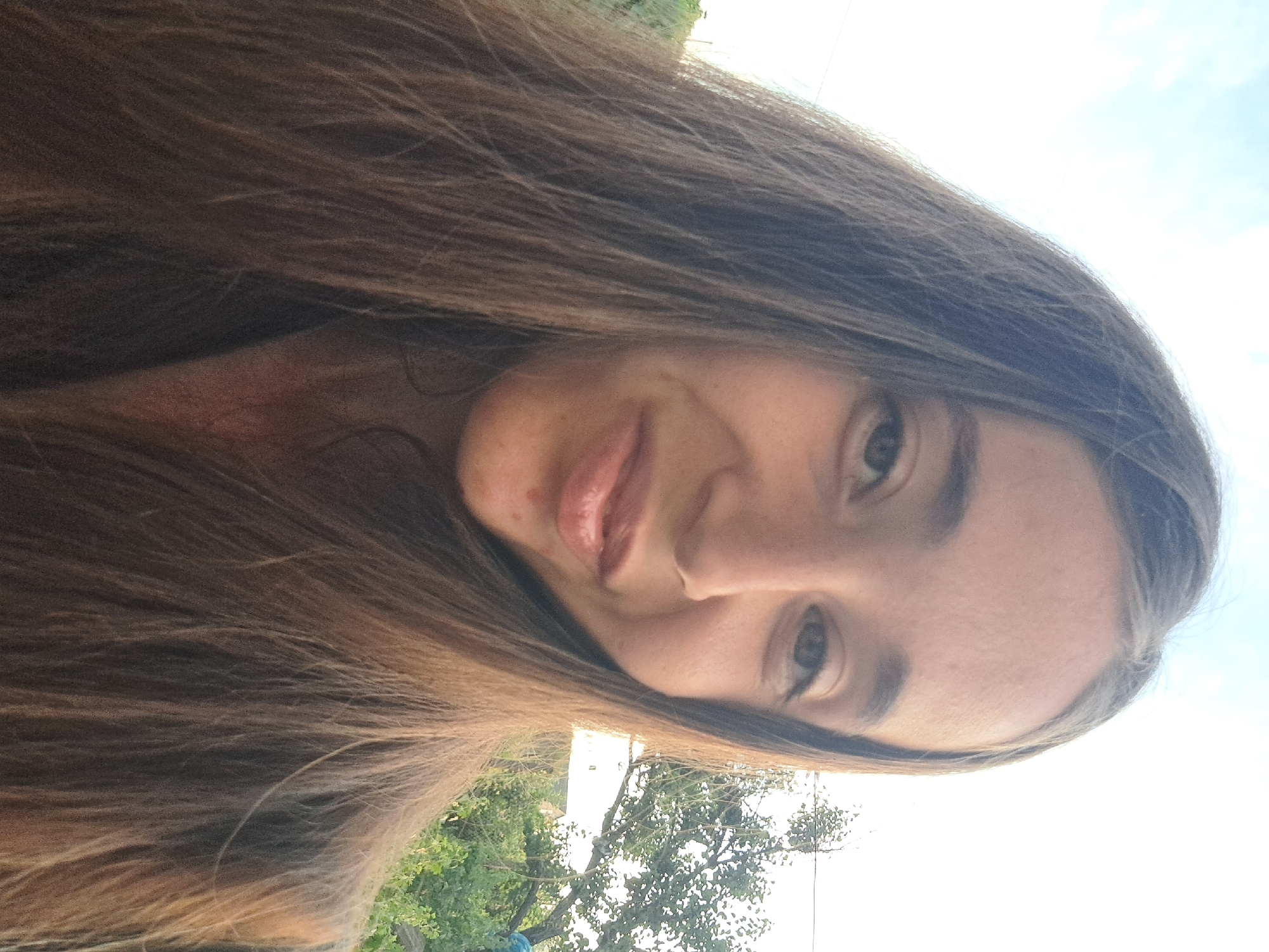 The image is a close-up, selfie-style photograph of a young woman with long, brown hair, taken outdoors. She is looking towards the camera with a slight smile. Her hair frames her face, and the background shows greenery and trees, suggesting a rural or suburban setting. The sky is visible in the upper portion of the image. The specific location in Vypasne, Ukraine, cannot be determined from the visual information provided in the image itself.
