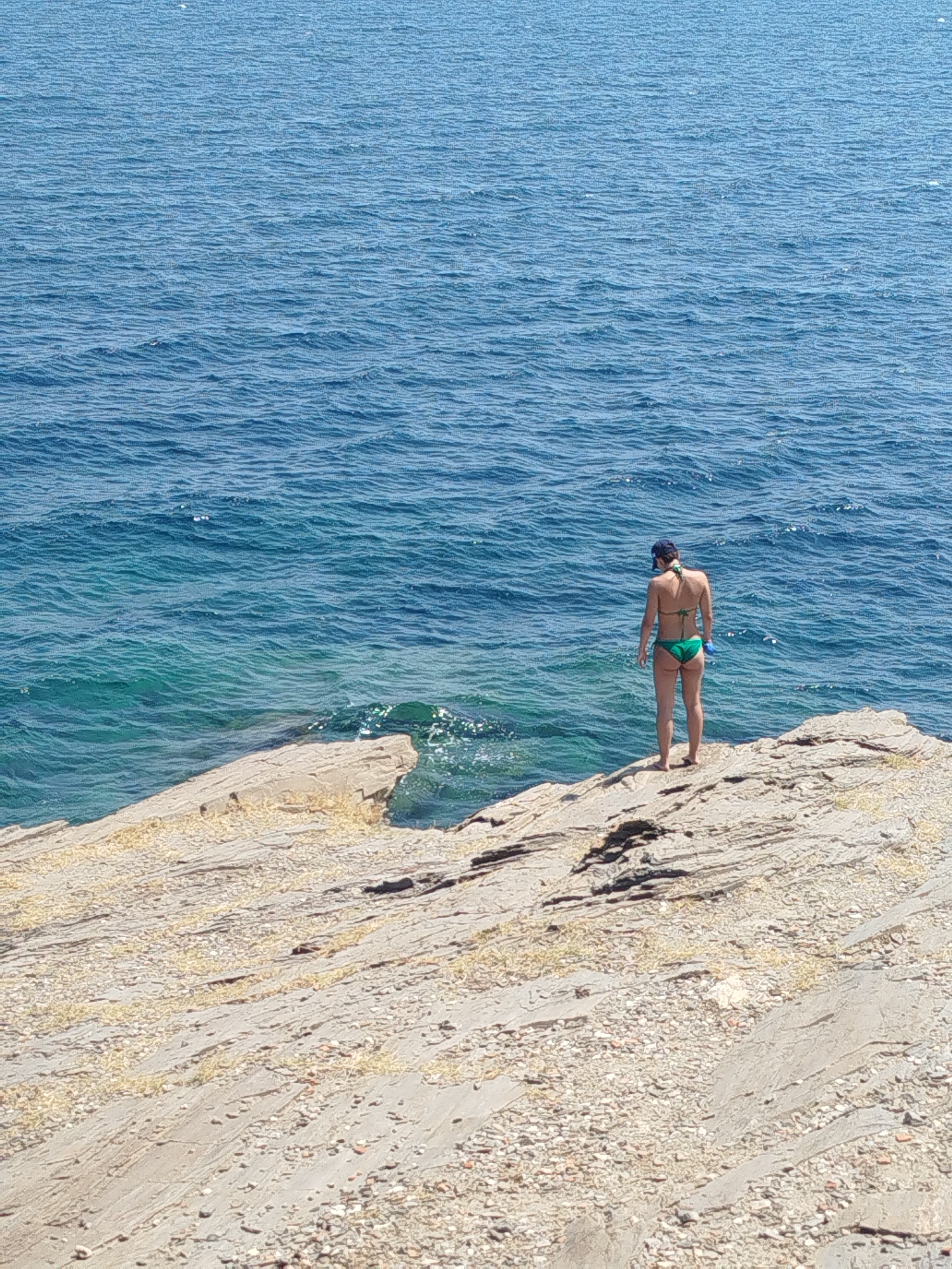 An adult woman stands on a rocky outcrop, facing the ocean. She is wearing a green bikini and a dark baseball cap. The sea is a deep blue with lighter turquoise hues near the shore. The rocks are layered and textured, with sparse vegetation. The scene suggests a coastal environment in Sithonia, Greece.