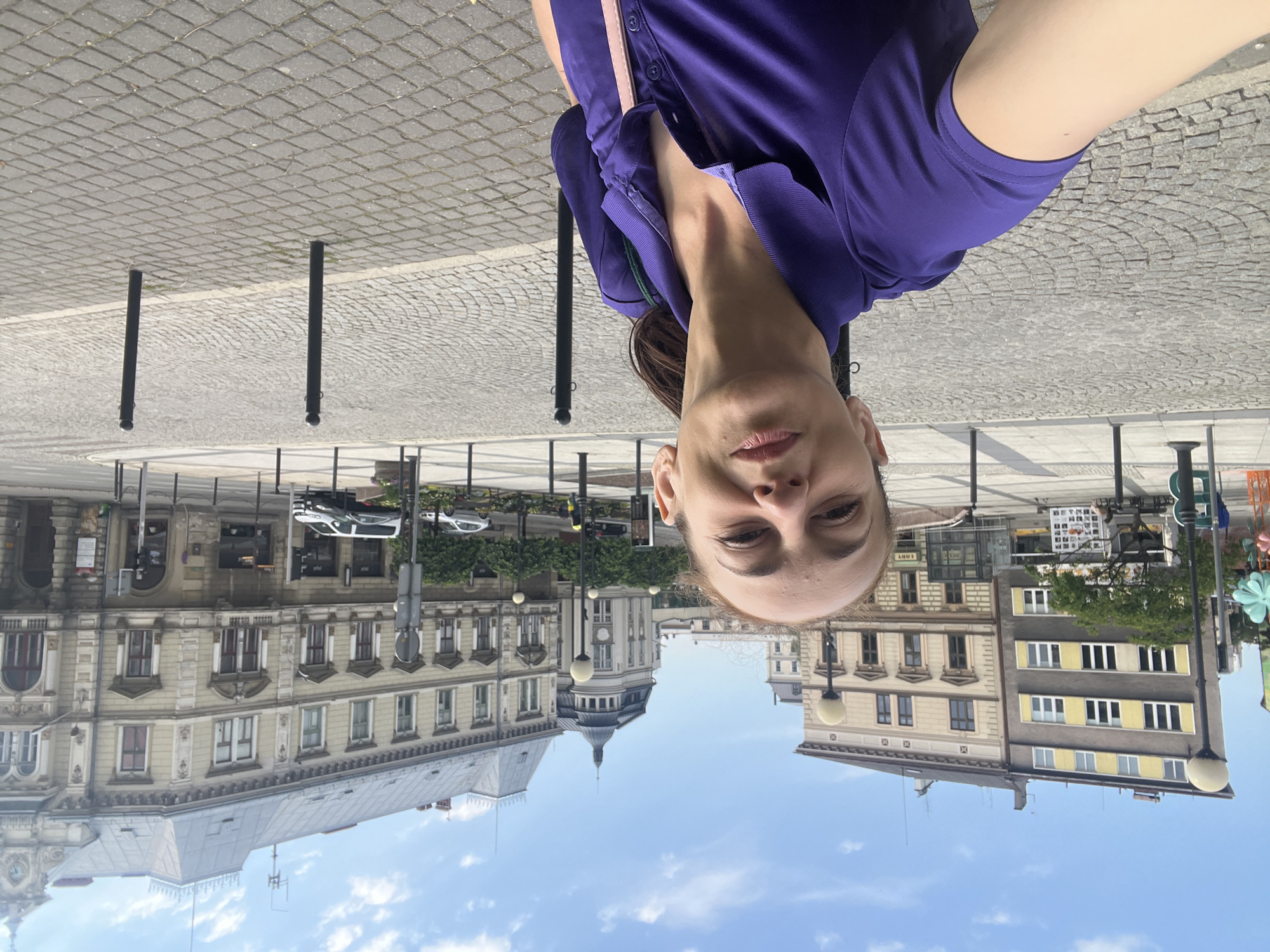 A woman with auburn hair tied back in a ponytail takes a selfie in a European city square. She is wearing a purple polo shirt and a pink strap is visible across her chest. Behind her, cobblestone pavement leads to a street with parked cars and a line of trees. Various buildings with traditional architecture, including a light-colored building with ornate details and a darker building with yellow window shutters, are visible in the background. The sky is blue with scattered clouds.