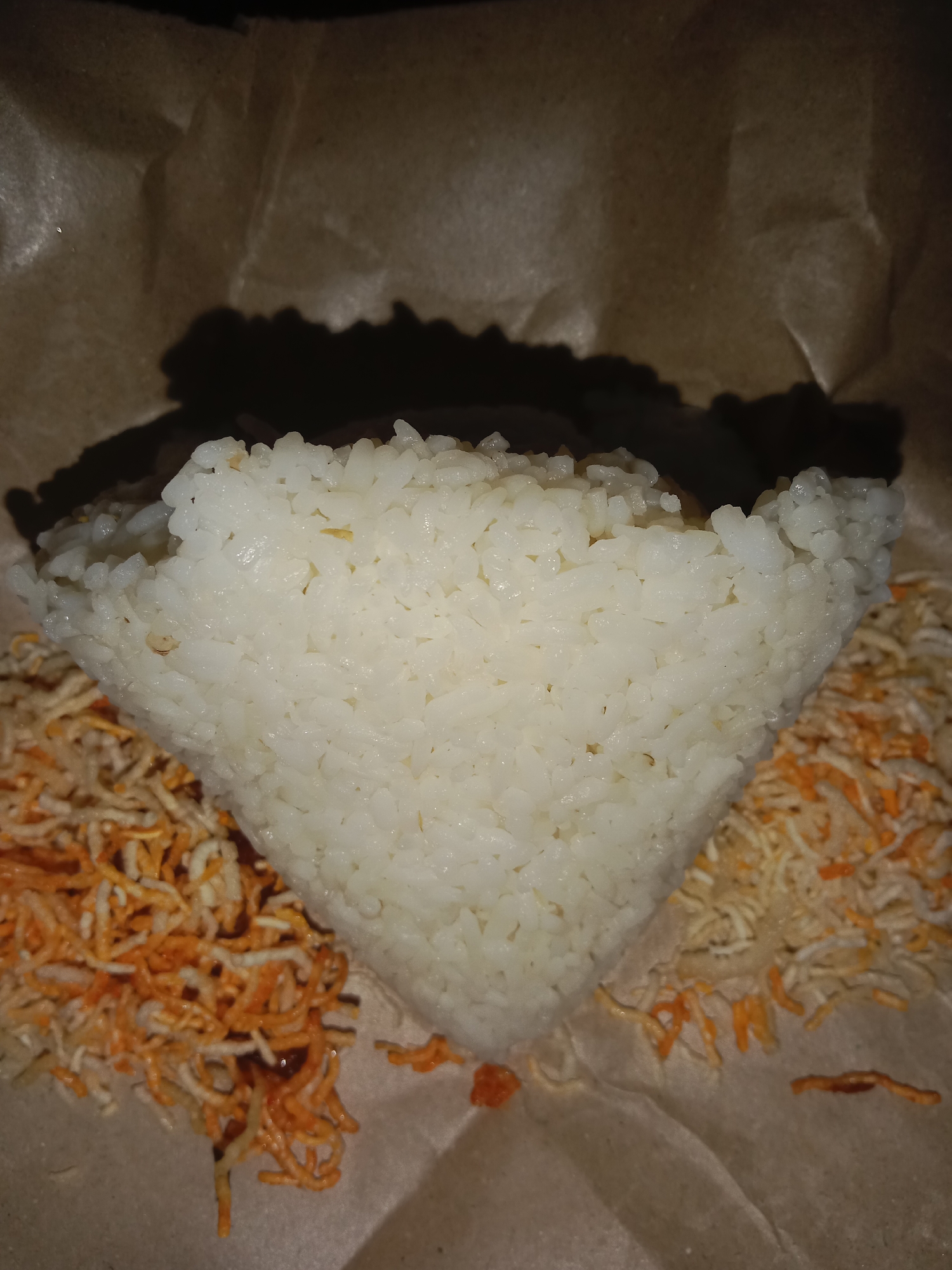 A close-up, overhead shot shows a portion of white rice forming a pyramid shape, positioned on top of brown paper. To the left and right of the rice are piles of thin, shredded orange and white food items, possibly noodles or shredded vegetables. The lighting casts a shadow on the paper behind the rice, creating a dark, indistinct shape. The scene appears to be set on a table, with no other objects or individuals visible. The location is the city of Pohsangit Leres, Indonesia.