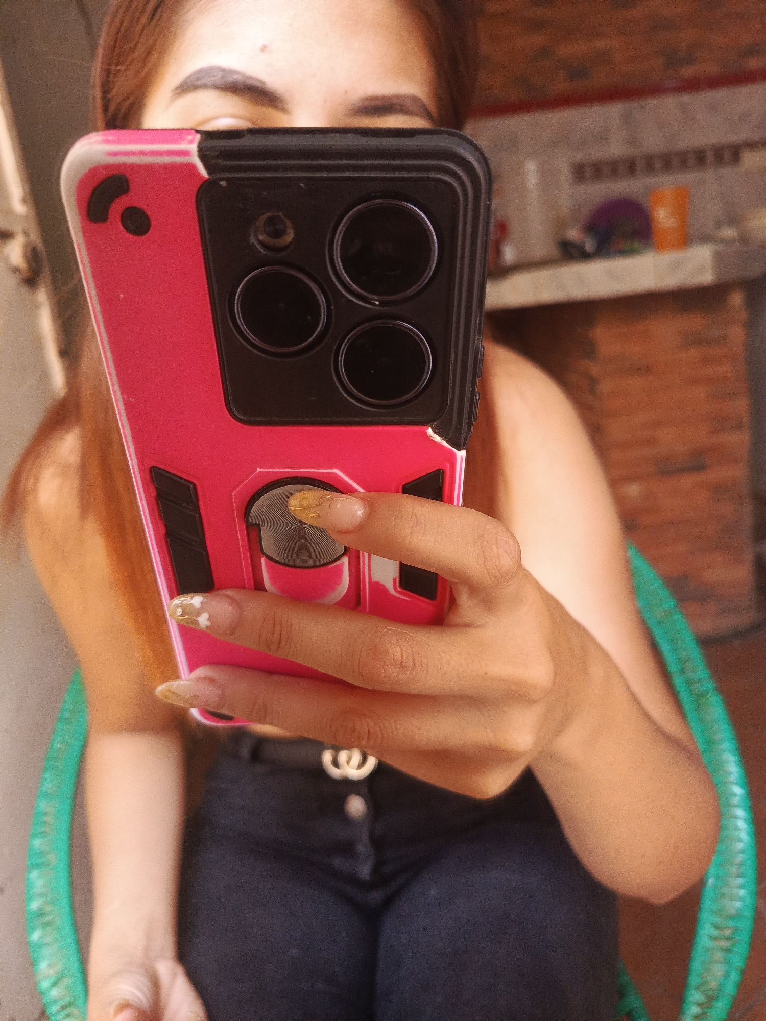 A person is holding a pink and black cell phone up to their face, obscuring the lower half of their face from the eyes down. Only the eyebrows and a small portion of the forehead are visible. The person has long brown hair. The phone has three lenses. The person is sitting in a green plastic chair, wearing a black shirt and black pants with a silver belt buckle. The background appears to be an indoor setting with a brick-like wall and a countertop with items on it. Based on the image context, the location is assumed to be Juan Griego, Venezuela.