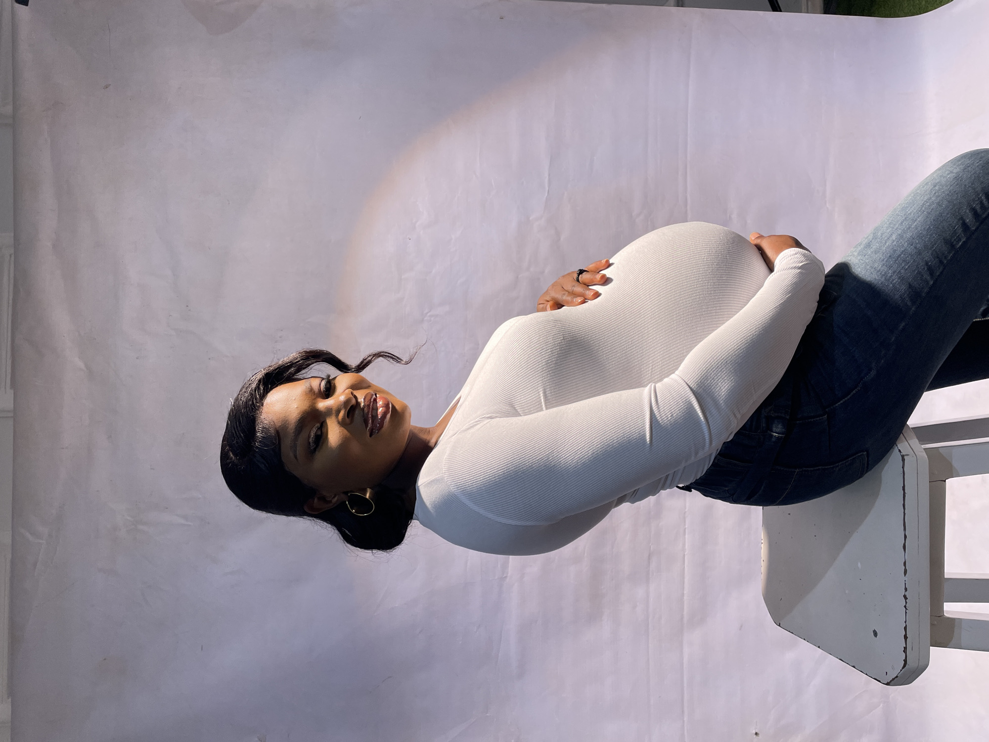 A pregnant woman with brown skin and long dark hair, wearing a white long-sleeved shirt and blue jeans, is seated on a white chair against a white backdrop. The woman is looking at the camera and holding her stomach. The location is Jos, Nigeria.