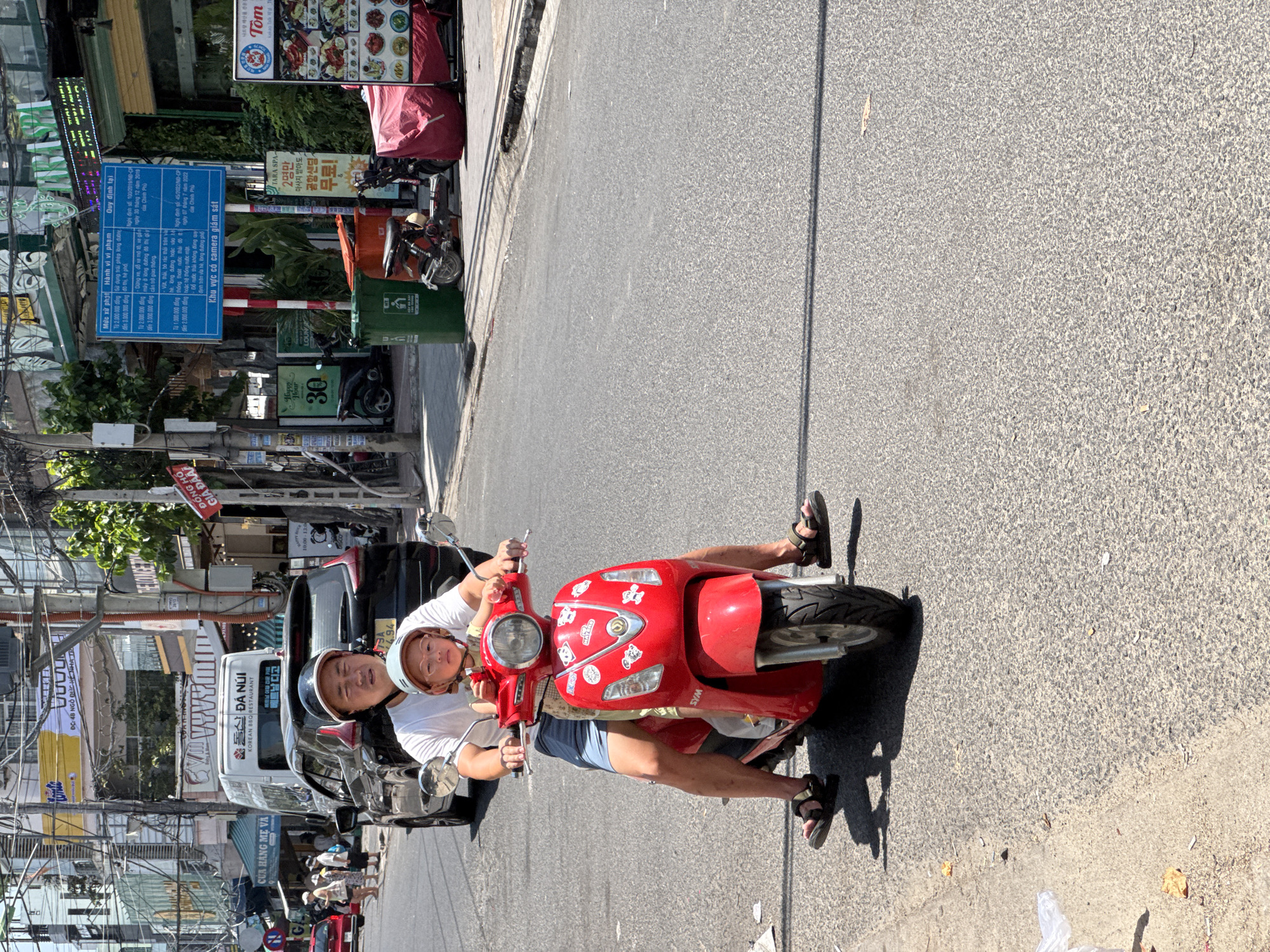 In Nha Trang, Vietnam, a man and a young child ride a red scooter on a sunny day. The man, wearing a white t-shirt and blue shorts, drives the scooter with the child, who wears glasses and a light-colored top, seated in front of him. Both are wearing helmets. The scooter is adorned with various stickers, including cartoon characters. They are on a street with buildings lining the left side, featuring shops and signage in Vietnamese and possibly other languages. A black SUV is visible behind them, along with other vehicles and parked motorcycles further down the street. The road appears to be asphalt, with some debris and shadows cast on its surface. A blue sign with Vietnamese text is posted on a pole to the right, indicating local regulations.