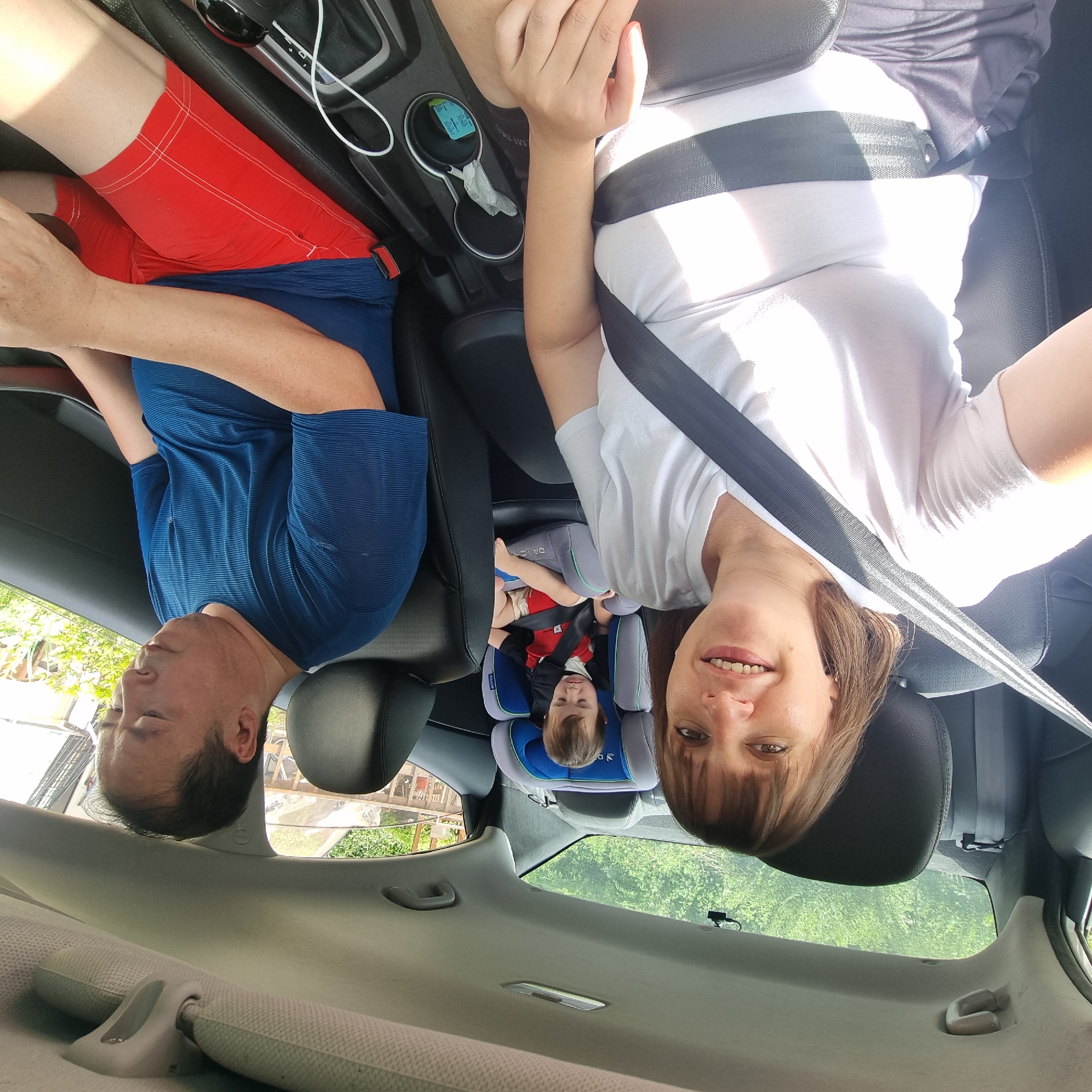 In Dunnae-myeon, South Korea, a family of three is seated in a car. A woman is in the front passenger seat, looking towards the camera with a smile. She is wearing a white t-shirt and has a seatbelt fastened across her chest. Next to her, in the driver's seat, is a man wearing a blue t-shirt and red shorts. He is looking down. In the back seat, a young child is secured in a car seat, dressed in a red and black shirt and red shorts. The car's interior is visible, including the seats, dashboard, and cup holders. Outside the car, greenery and trees can be seen through the windows.