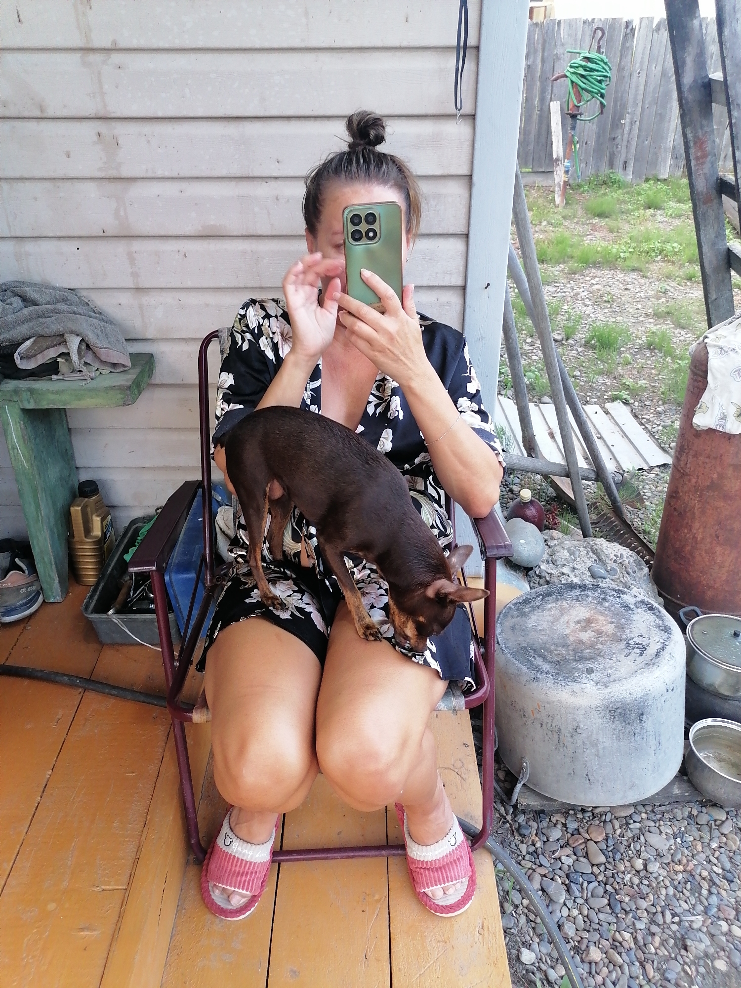 A dark-haired woman with her hair in a bun is sitting in a folding chair on a wooden porch. She is wearing a black and floral patterned dress and pink and white striped slippers. The woman is holding a green smartphone in her right hand, obscuring her face as she appears to be taking a selfie. A small, dark brown dog is standing on her lap, with its head facing down. To the left of the woman, on a green shelf, are stacked grey towels and various containers. Further to the left and below the shelf, a pair of blue and grey sneakers are visible. The porch has a weathered wooden floor. Behind the woman, a wooden fence and some vegetation are visible. To her right, there are several large metal pots and a rusty vertical object. The scene appears to be outdoors in a rural or semi-rural setting, possibly in Novopokrovka, Russia, given the typical structures and environmental elements.