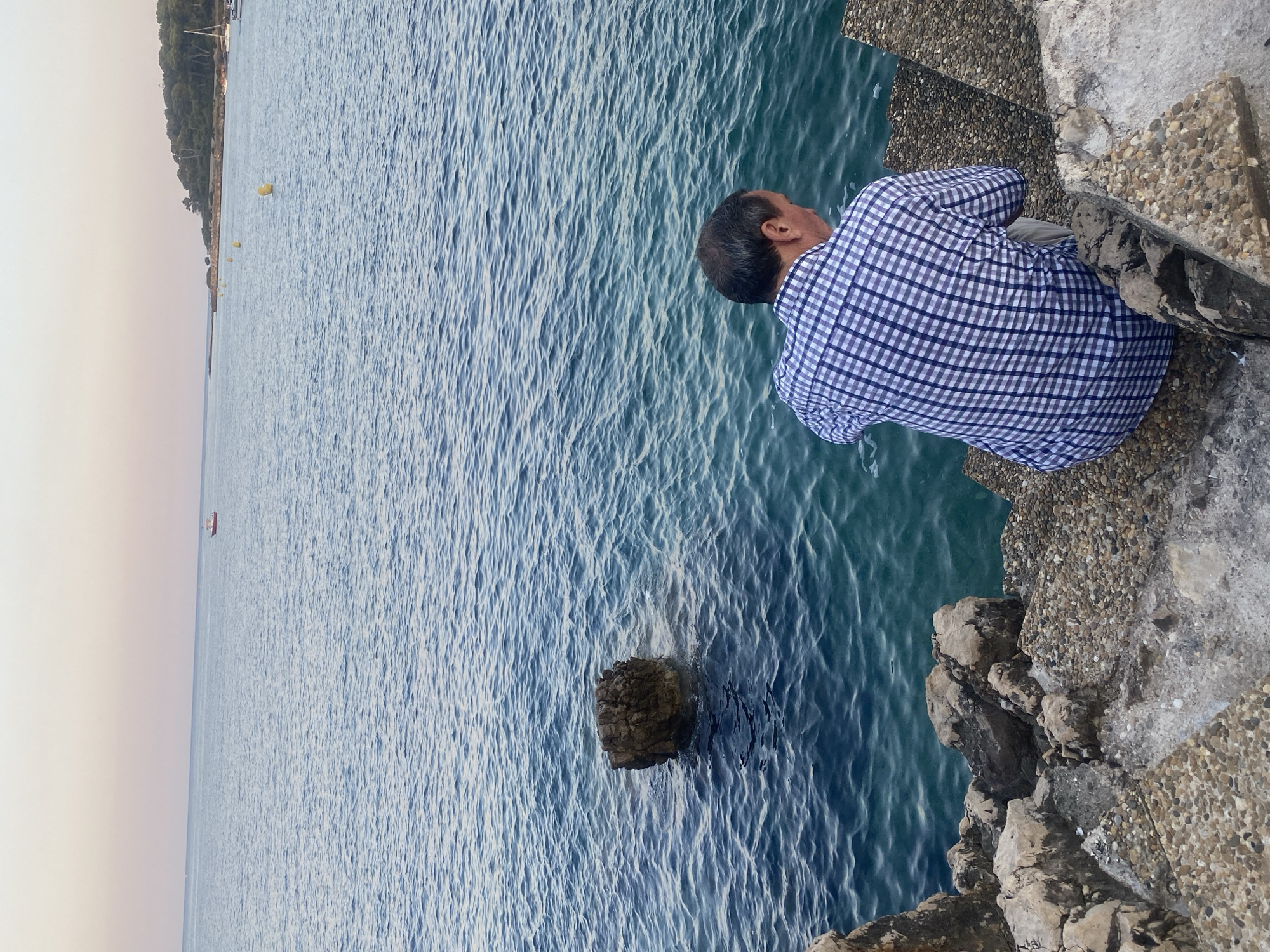 The image depicts a man wearing a blue and white checkered shirt and beige pants sitting on a stone staircase facing the sea. The sea is a vibrant blue, with gentle waves. In the distance, a small red boat is visible on the water. The coastline, with trees and some structures, can be seen further away, under a pale sky. The scene appears to be a waterfront area in Antibes, France, possibly near a harbor or a rocky shore. The man is positioned on the right side of the frame, looking down towards the water.