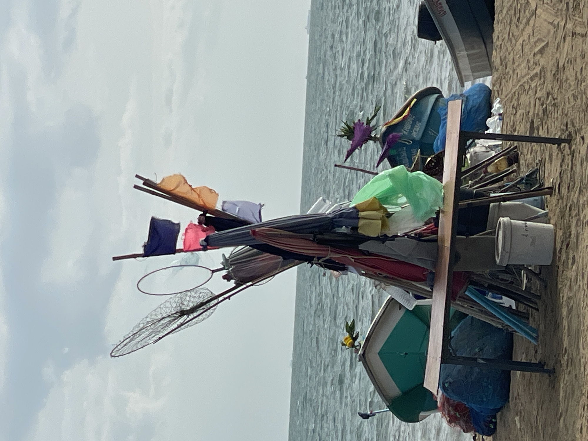 The image depicts a beach scene in Pattaya City, Thailand. In the foreground, a rustic wooden table with metal legs stands on the sand. To the left of the table, a green and white boat is partially visible, adorned with a yellow flower. A cluster of fishing nets and assorted items, including flags and umbrellas, are piled and propped up on wooden poles in the center of the frame. Several fishing boats are also present, some partially submerged in the water. The sea occupies the background, with a hazy horizon. The sky is overcast with clouds.
