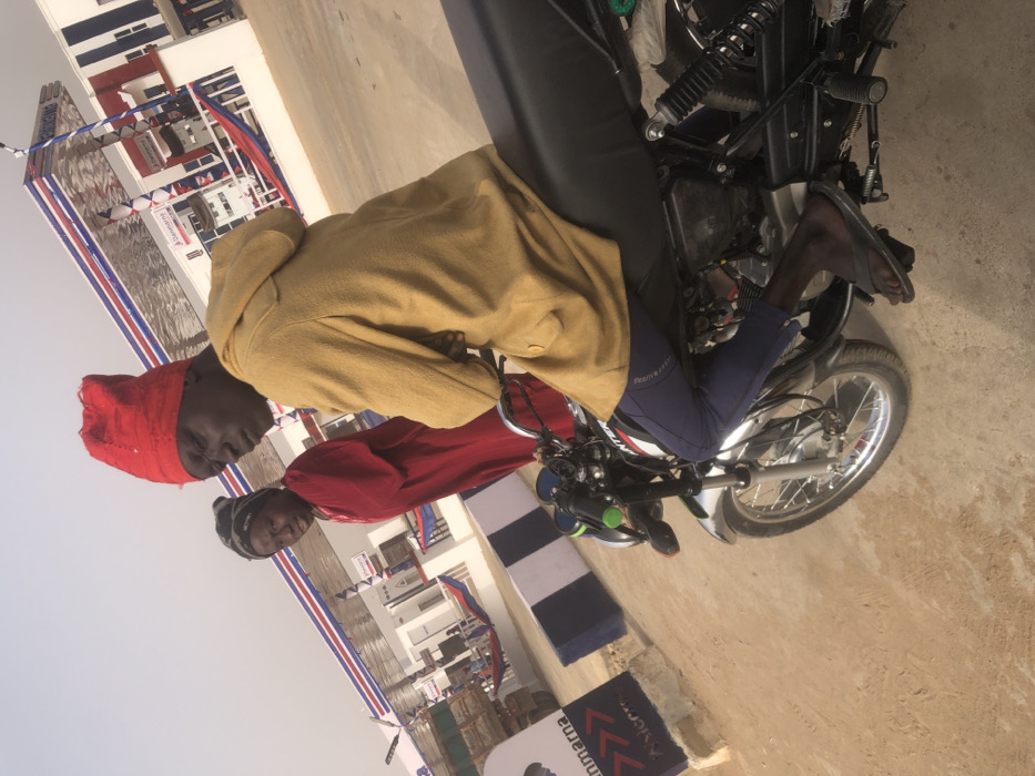 Two men stand near a motorcycle. One man wears a red head covering and red shirt, while the other wears a tan jacket. They are positioned in front of a building decorated with banners and flags in red, white, and blue. The motorcycle is black with silver wheels. The ground is light brown and appears to be a sandy or dusty surface. The sky is overcast.