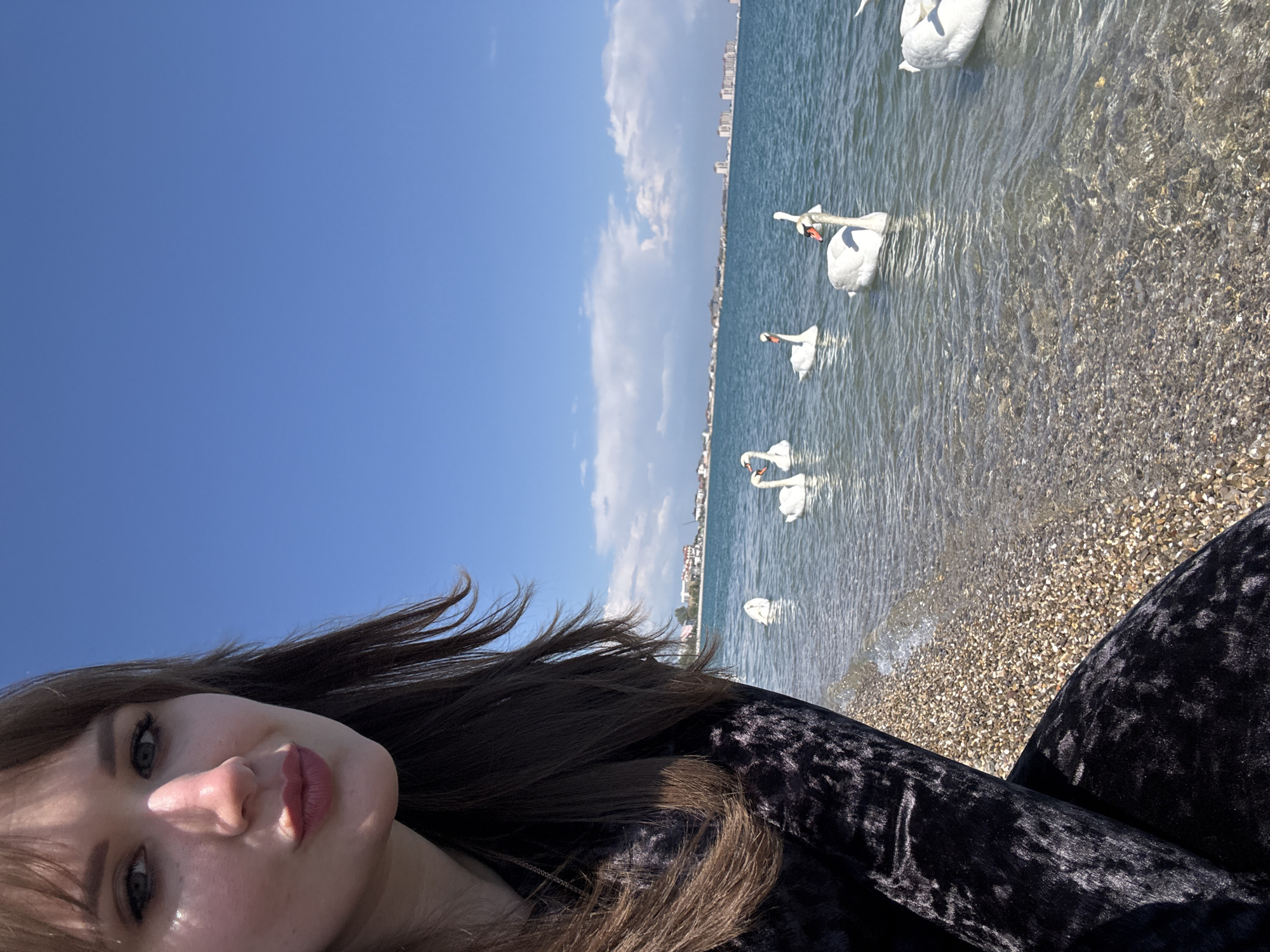 The photograph depicts a woman, possibly taking a selfie, with a beach scene as the backdrop. The location appears to be in Yevpatoriya, Ukraine, as evidenced by the coastal buildings in the background. Several swans are visible in the water near the shore. The water is clear, revealing a pebble beach. The sky is blue with some clouds. The woman has brown hair and is wearing a dark velvet-like garment.