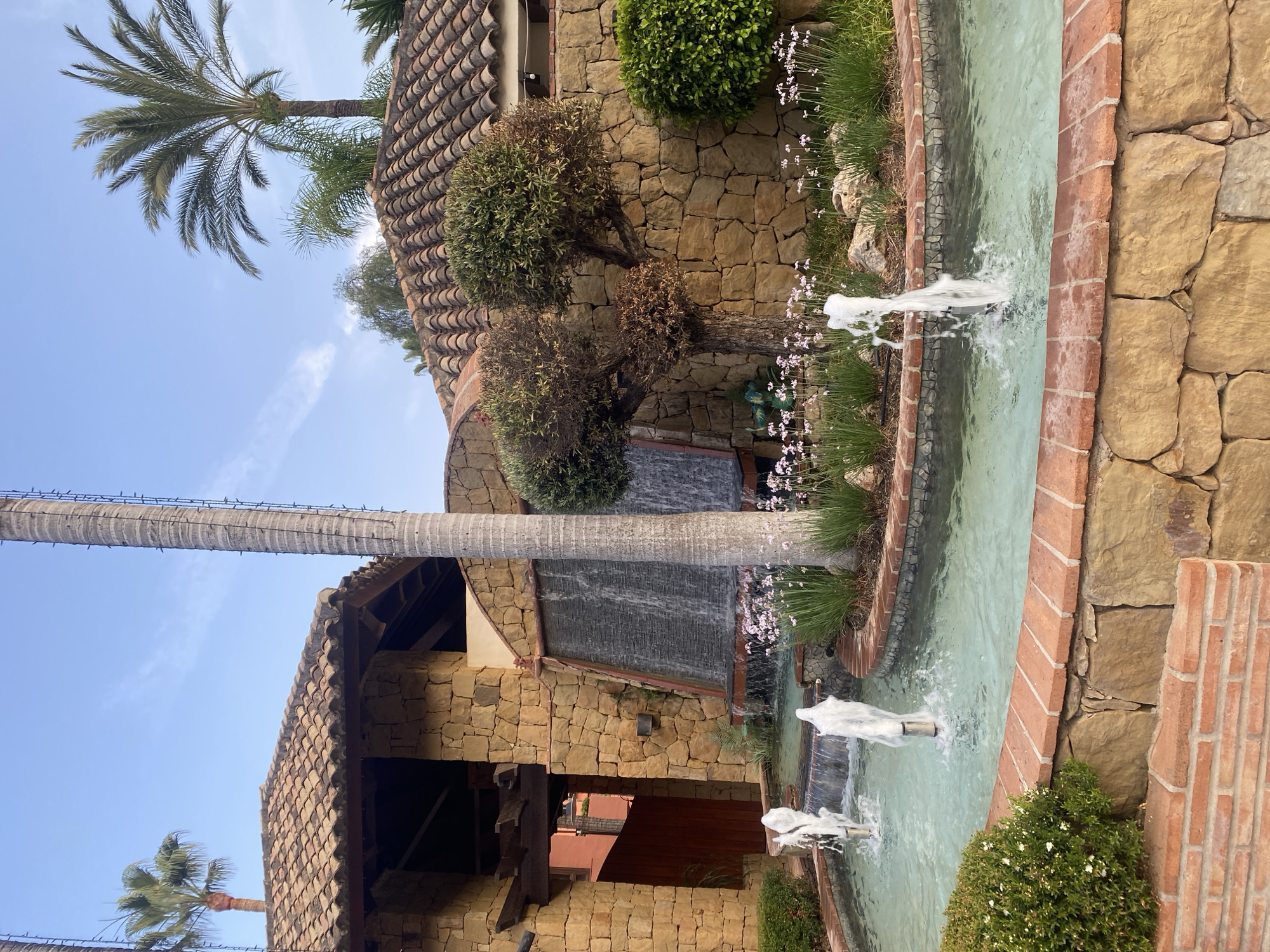 The image shows a man-made water feature with multiple fountains and a cascading waterfall. The architecture appears to be of Mediterranean or Spanish style, with stone walls and terracotta tiled roofs. Lush greenery, including palm trees and various shrubs and flowers, surrounds the water feature, suggesting a warm climate. The lighting indicates it is daytime, possibly late afternoon given the soft light. The scene is tranquil and suggests a well-maintained outdoor space, likely a resort, hotel, or private residence in a coastal town. Without additional information or context, pinpointing the exact location within Estepona, Spain, is impossible, but the architectural style and vegetation are consistent with the region. There are no people visible in the image.