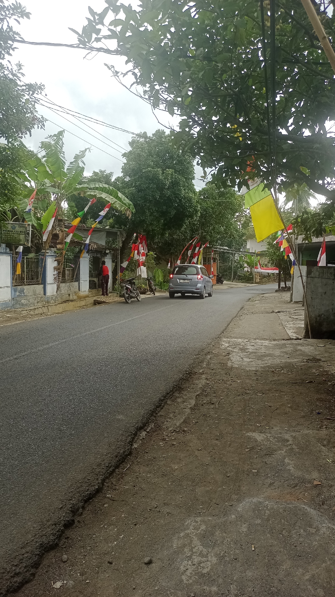 The image depicts a street scene in Cibeber, Indonesia, decorated with numerous red and white Indonesian flags strung across the road and along the side of the buildings. A light-colored car is driving away from the camera down the asphalt road, which has a rough edge on the right side where it meets the dirt shoulder. Two motorcycles are parked on the side of the road near a building on the left. A person wearing a red shirt is standing near the building. Trees with green foliage are prominent throughout the scene, and the sky is overcast. The buildings appear to be residential or small commercial establishments.
