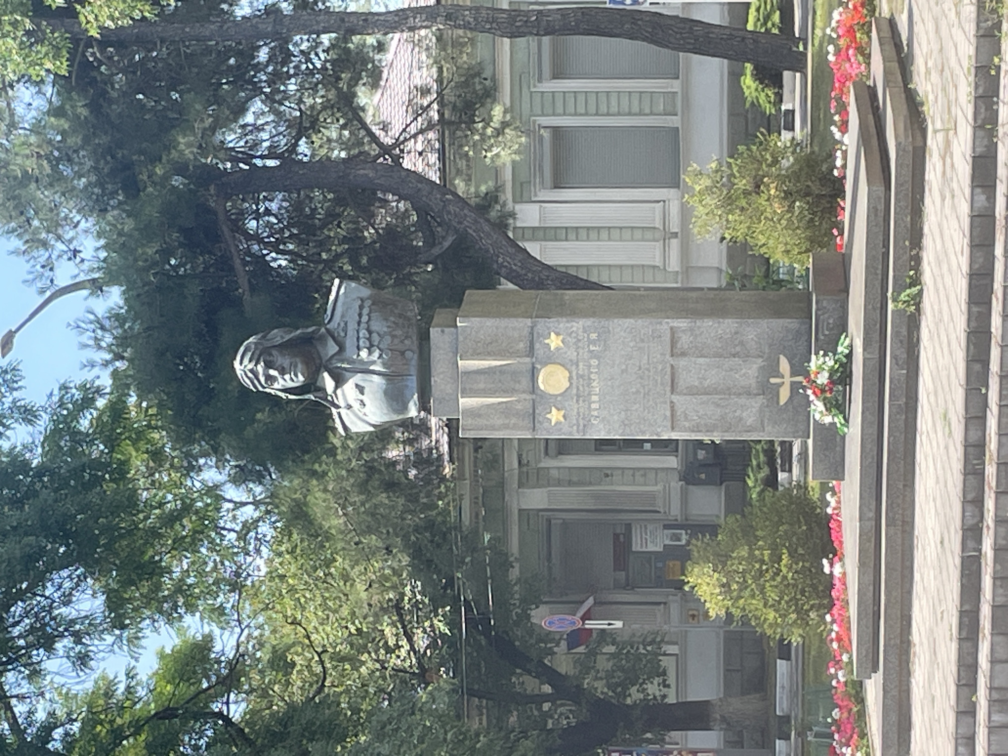 The image shows a monument in Novorossiysk, Russia, dedicated to Hero of the Soviet Union Valery Serkov. The monument features a bronze bust of Serkov in a military uniform, mounted on a granite pedestal. The pedestal is adorned with two stars and an inscription. In front of the pedestal, there is a wreath of flowers. The monument is surrounded by a paved area and a flower bed. In the background, there is a light-colored building with columns and windows, and several trees. The sky is visible above the trees, with some clouds. The overall scene depicts a public space with a memorial.