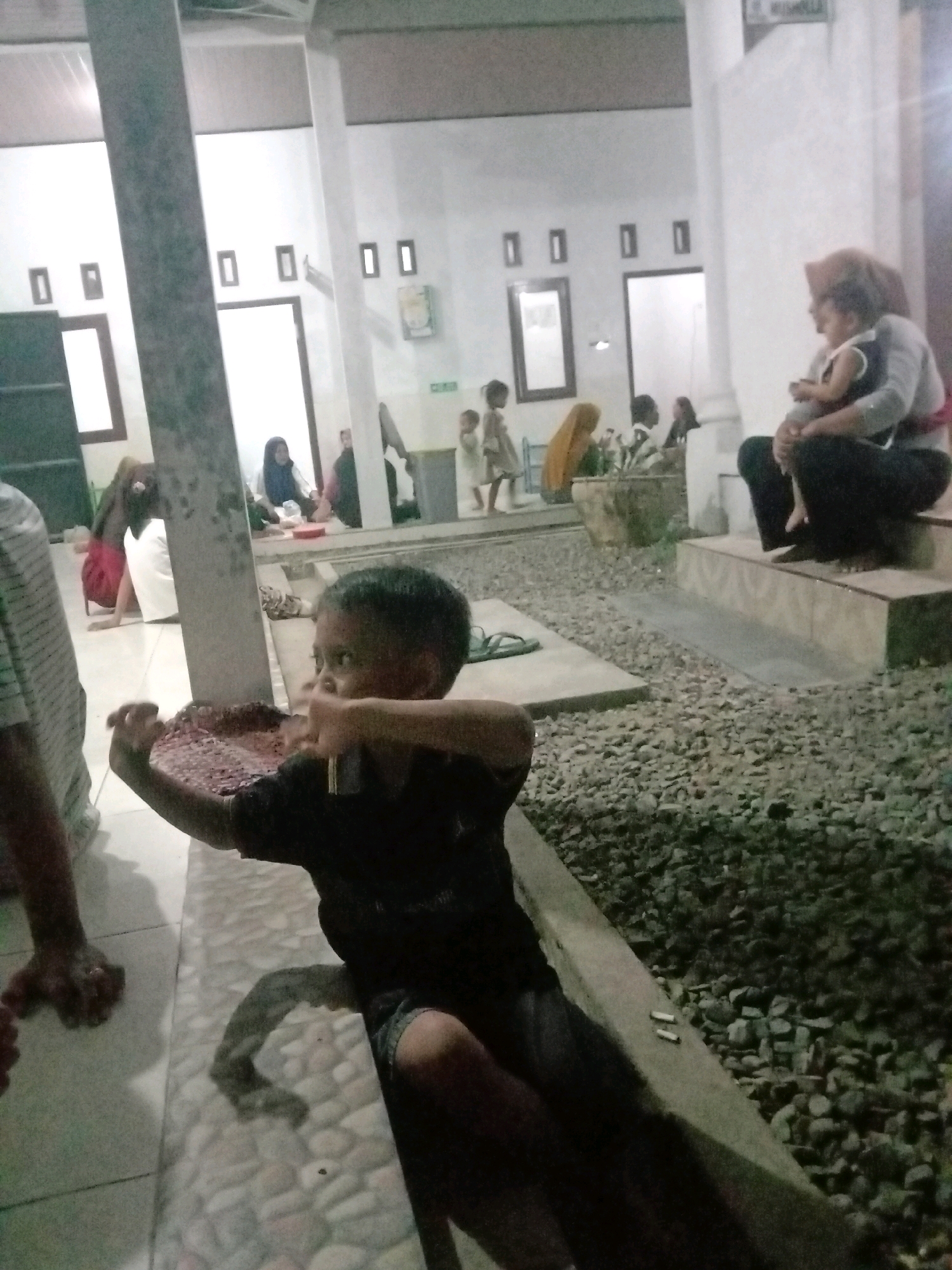 The image depicts a nighttime scene with low lighting, possibly an outdoor gathering area within a residential or community complex in Rejosari Mataram, Indonesia. In the foreground, a young boy, appearing to be around 5-7 years old, is in focus, actively engaged with a woven mat or tray. He is wearing a dark t-shirt and shorts. His facial expression is intent as he manipulates an object within the mat. To his left, partially visible, is the arm and torso of an adult wearing a striped shirt, suggesting they are nearby.
In the background, several other individuals are present, though less clearly defined due to the lighting and focus. On the right side, an adult woman, wearing a headscarf and dark clothing, is seated and holding a baby on her lap. The baby, wearing a white garment, is also visible. Other figures, including children and adults, are scattered throughout the background, some appearing to be seated on the ground or on steps. The environment consists of a paved area with small stones, common in Indonesian outdoor spaces. Structures with white walls and rectangular windows are visible, consistent with local architecture. The overall atmosphere appears to be a casual social gathering.