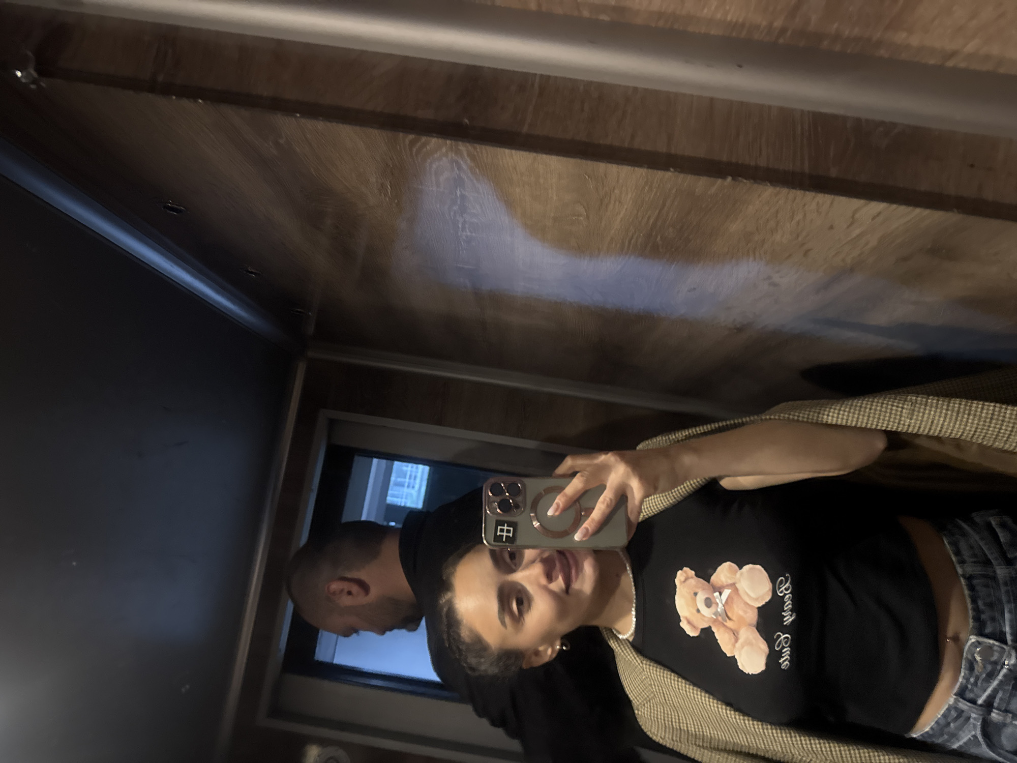 A selfie shows a woman taking a picture in an elevator with a man, presumably her partner, standing behind her. The woman has dark hair pulled back, wears a black t-shirt with a teddy bear graphic, blue jeans, and a tan blazer. She is holding an iPhone with a dark-colored case and a circular magnetic ring. The man behind her has a shaved head and wears a dark-colored top. The elevator interior appears to be made of wood paneling. The location is likely Tbilisi, Georgia, given the likely origin of the image.