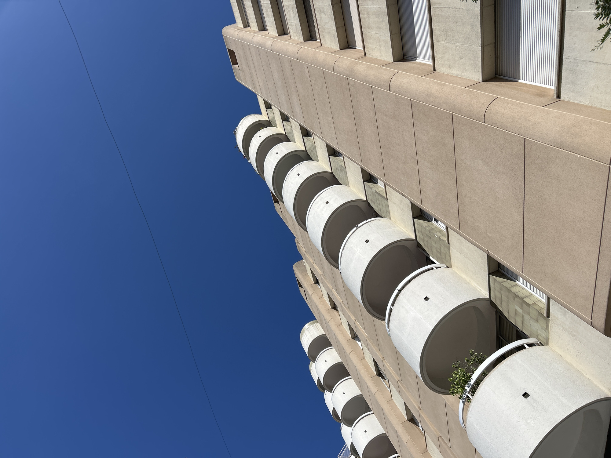 An upward angle shot captures the facade of a modern apartment building under a clear blue sky. The building's exterior is a muted beige, with cream-colored circular balconies protruding from its side. The balconies are stacked in a staggered pattern, creating a dynamic visual rhythm. Some of the balconies appear to have small plants growing in them. The sky above is a deep, uninterrupted blue, with a thin black wire stretching diagonally across it. The image is taken from the perspective of someone standing on the ground and looking up at the building. The scene is devoid of any people. The architecture suggests a contemporary urban environment, and the clean lines and geometric shapes are characteristic of modern design. The lighting indicates a bright, sunny day.