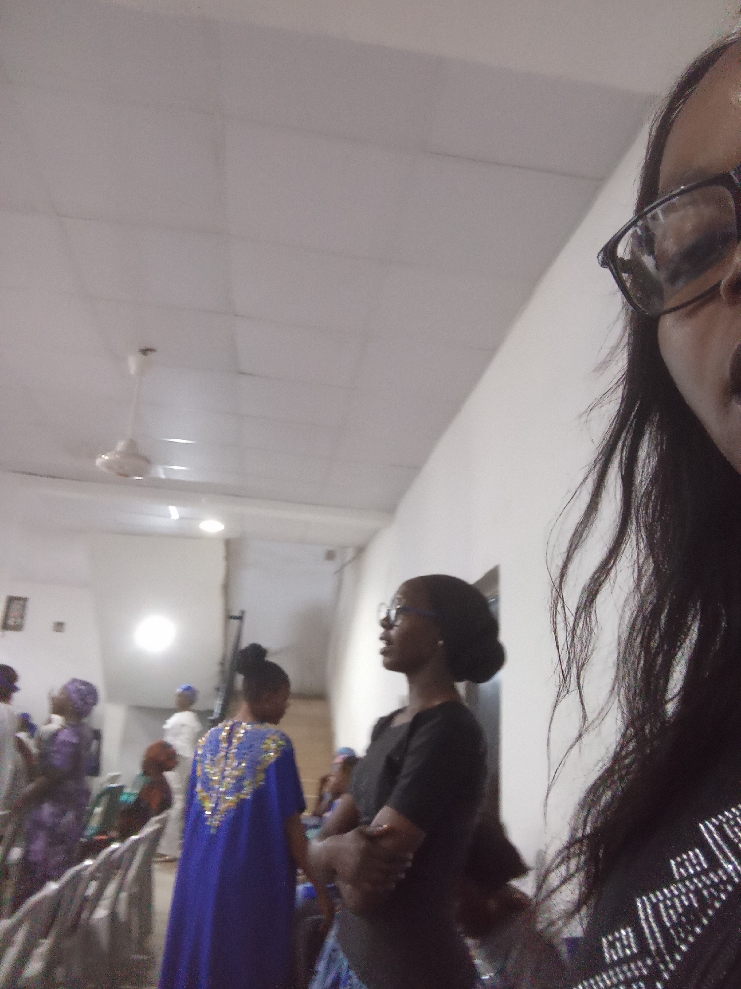 The image captures an indoor gathering, likely a religious service or event, in Kason, Nigeria. The scene is illuminated by overhead lighting and a ceiling fan is visible. In the foreground, a person wearing glasses and a dark top is partially visible on the right side of the frame, with their hair and upper body in focus. To the left and in the midground, several individuals are present. A woman in a vibrant blue dress adorned with gold embellishments stands facing away from the camera. Other attendees, dressed in various traditional Nigerian attire, are also visible, some seated and others standing. The environment appears to be a community hall or similar venue, with white walls and a tiled ceiling. The overall atmosphere suggests a formal occasion.