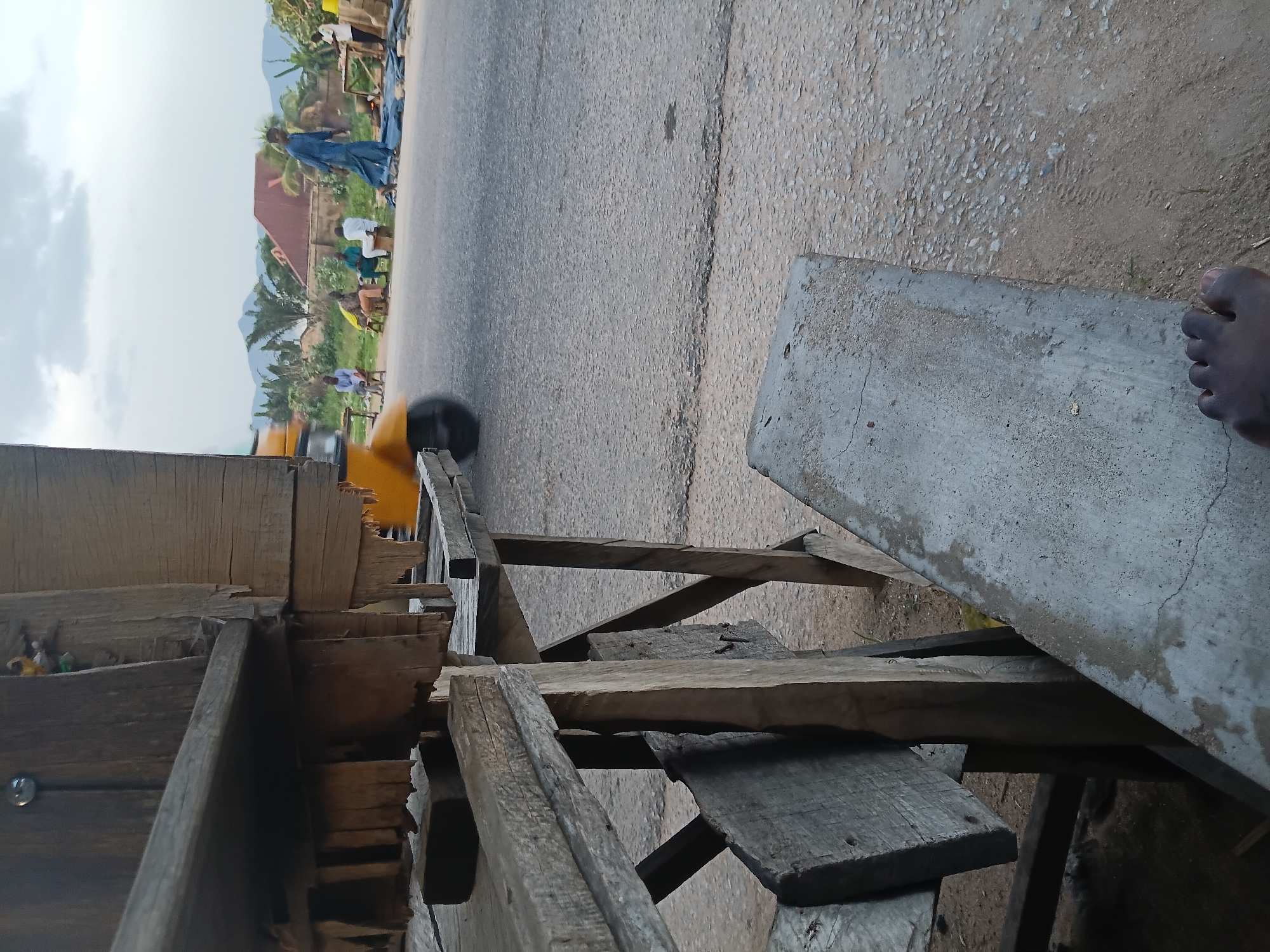 An eye-level, medium shot shows a roadside scene in Jalingo, Nigeria. In the foreground, weathered wooden structures, possibly a market stall or seating area, are prominent. A rough-hewn concrete bench is in the lower right, with a person's bare foot resting on it. In the midground, a dusty asphalt road runs horizontally. A yellow auto-rickshaw is partially visible on the left, moving away from the camera. Several individuals are scattered along the roadside in the midground. One man in a blue robe walks away from the camera on the right side of the road. Other men, dressed in casual attire, sit on stools or the ground near the road. Some appear to be interacting, while others are seated individually. The background features a line of trees, houses with reddish roofs, and distant mountains under a cloudy sky. The overall atmosphere suggests a quiet moment in a semi-urban or rural setting.