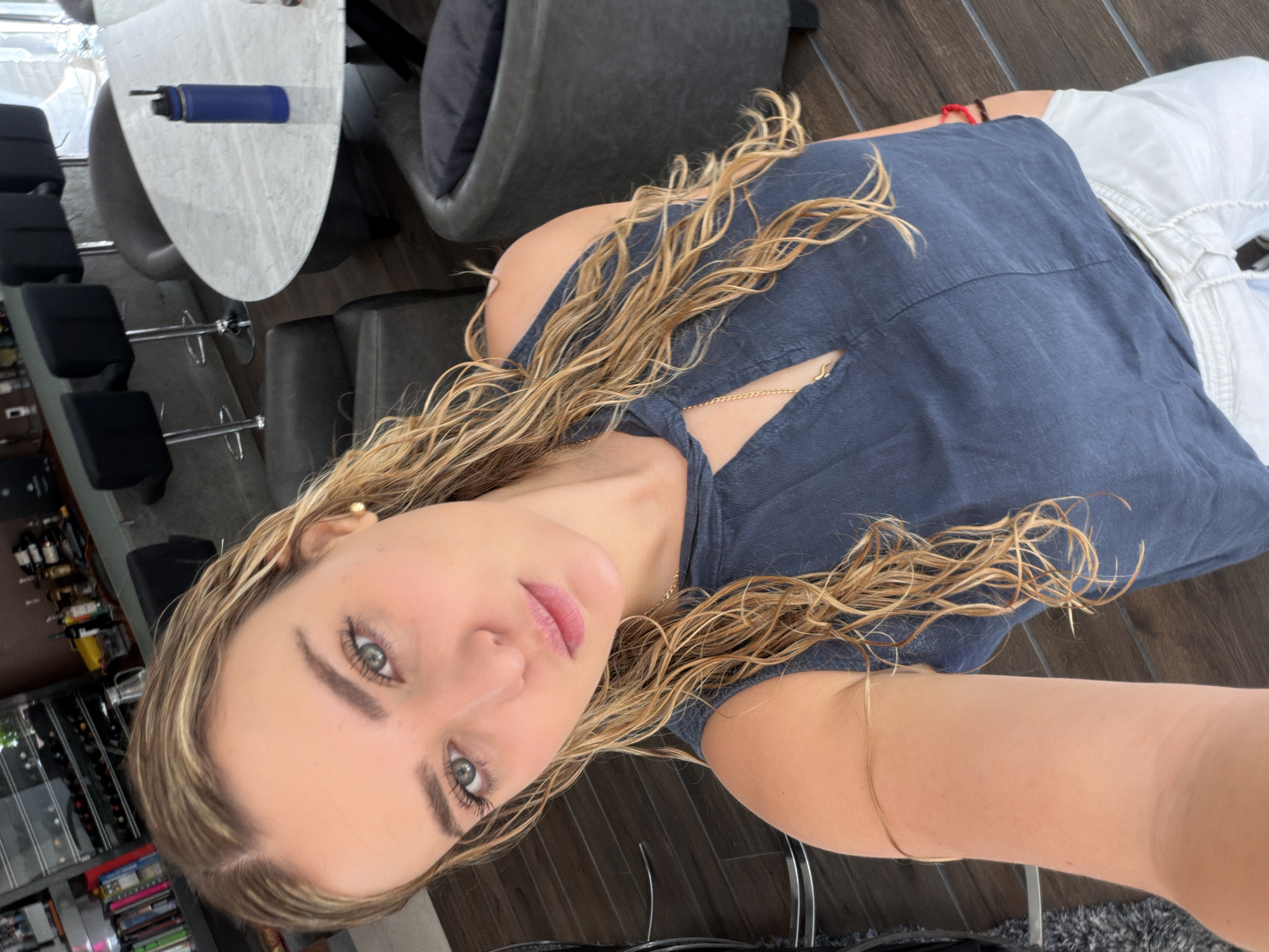 A female subject is captured in a close-up selfie indoors, located in San Bernardino Tlaxcalancingo, Mexico. The subject has fair skin, light green eyes, and long, wavy blonde hair that appears damp. She is looking directly at the camera with a neutral expression. She wears a dark blue sleeveless top with a V-neck and a small cut-out, a thin gold chain necklace with a small pendant, white pants with a drawstring waist, a thin gold-colored bangle on her left arm, and a red bracelet on her right wrist.
The background shows a modern interior space with dark wooden flooring. To the left, a dark shelving unit containing bottles and books is partially visible, along with three black bar stools with chrome bases. To the right, a white oval table with a textured surface holds a blue insulated water bottle. Dark grey upholstered chairs, one with a dark pillow, are arranged around the table. Dark grey armchairs are also partially visible.