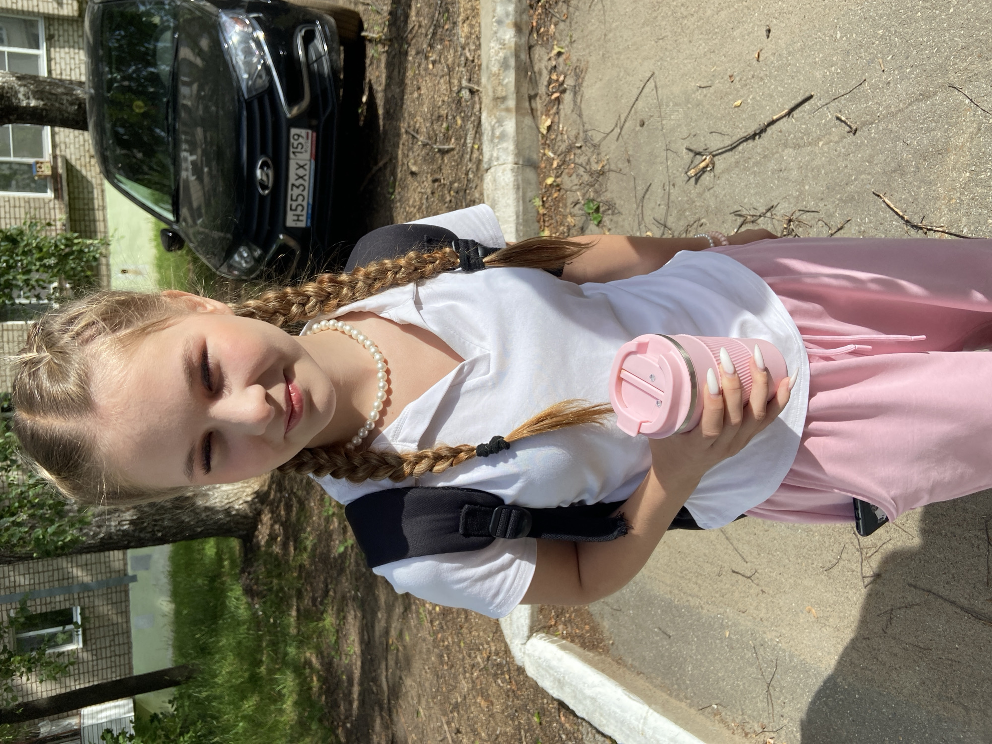 The image captures a young girl, likely a teenager, standing outdoors in Chaikovsky, Russia. She is wearing a white t-shirt, pink sweatpants, and a black backpack. Her blonde hair is styled in two thick braids. Around her neck, she wears a pearl necklace, and she has a pink portable blender in her right hand. Her fingernails are painted white.
The background shows a paved area, possibly a sidewalk or path, with dry leaves and twigs scattered on the ground. To the left, a black car with a license plate reading "H553XX 159" is parked, and behind it, a brick building with windows is visible, partially obscured by a tree.
The lighting suggests it is daytime, likely sunny, given the strong shadows. The girl's eyes are closed and she has a gentle smile, conveying a sense of contentment or perhaps a moment of quiet enjoyment. The scene appears to be a casual, everyday setting, possibly on the way to or from school, or during a break.