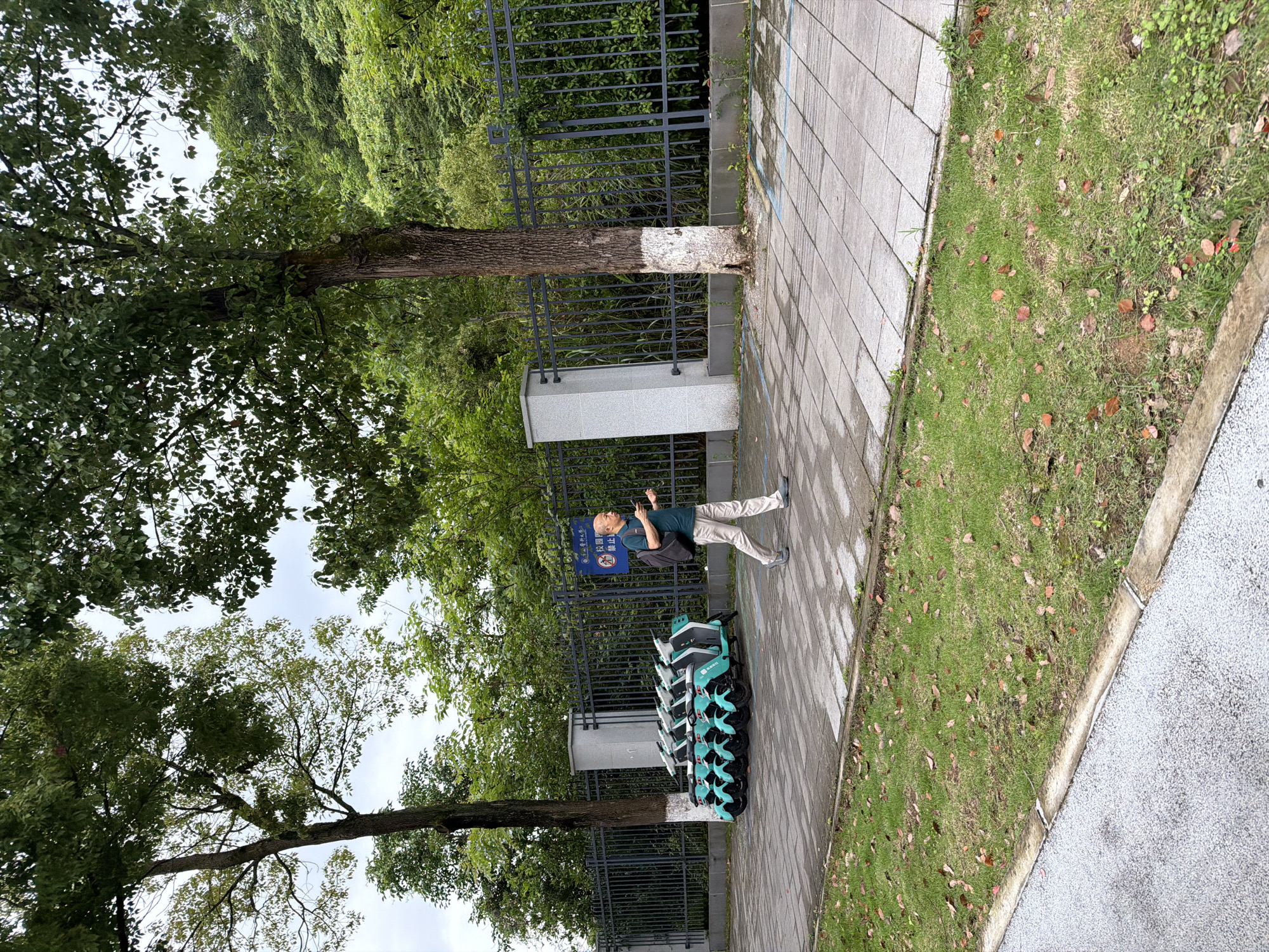 A photograph, taken outdoors in daylight, likely in the Chongqing High-tech Zone of China, shows a scene along a paved walkway. A middle-aged man is standing on the walkway, holding a small device. He is wearing a dark green shirt, light-colored pants, and grey shoes, with a black shoulder bag. To his left, a row of light teal and white electric scooters are neatly parked. The walkway is bordered by a black metal fence, behind which are trees and greenery. A tree trunk, partially painted white, stands near the fence. The sky is overcast. A patch of grass with fallen leaves is visible next to the walkway. The perspective suggests the photograph was taken from a slightly elevated position. A sign with Chinese characters is visible near the parked scooters.