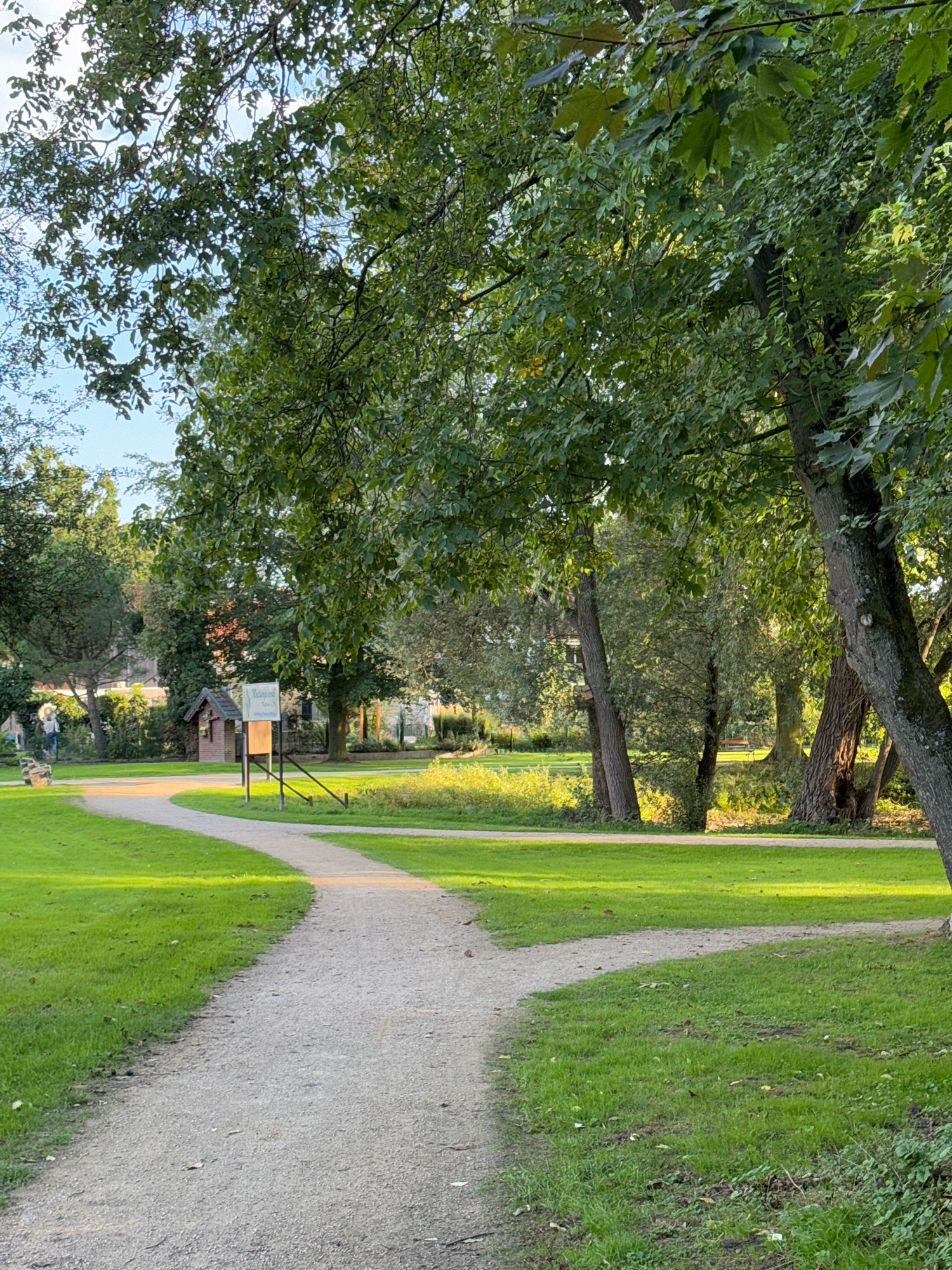 The image depicts a well-maintained park with a gravel path winding through lush green grass. Tall trees with dense foliage provide shade, dappling the scene with sunlight. In the background, a small wooden structure, possibly a playhouse or information booth, stands near a sign. Further in the distance, several buildings with red-tiled roofs are visible, suggesting a residential area adjacent to the park. A person can be seen walking in the far left of the image. The overall atmosphere is peaceful and serene, indicative of a public park. The location is identified as the park in Cleves, Germany.
