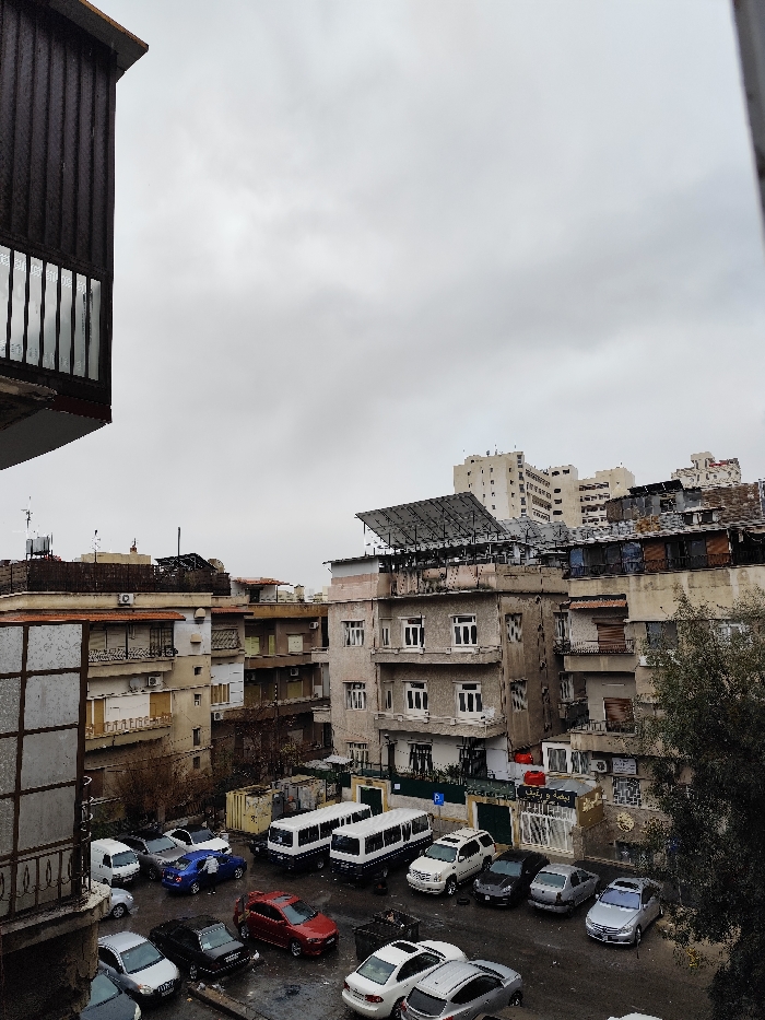 The image presents an urban landscape within As-Salihiya Municipality, Syria, under an overcast sky. The perspective is elevated, with a dark, vertically-slatted architectural element, likely a balcony or building exterior, partially framing the left foreground.
The mid-ground is dominated by a cluster of multi-story buildings exhibiting various architectural styles and states of repair. A prominent grey-toned building in the center-right features multiple windows and balconies, and its rooftop hosts a large, grid-mounted solar panel array. Other building rooftops display equipment such as water tanks, satellite dishes, and various utilities.
Below the buildings, an open-air parking area occupies the lower central portion of the image. This area contains numerous parked vehicles, including sedans of various colors (e.g., red, blue, silver, black), a white SUV, and two distinct white minibuses. A single individual is observed leaning over the front of a blue sedan in the parking area, suggesting an interaction with the vehicle. The ground surface of the parking area appears wet. Several dark-colored refuse bins are also visible within the parking zone.
To the right of the parking area, a tree with sparse foliage is present. In the distant background, taller, lighter-colored multi-story buildings are discernible against the grey, cloudy sky.