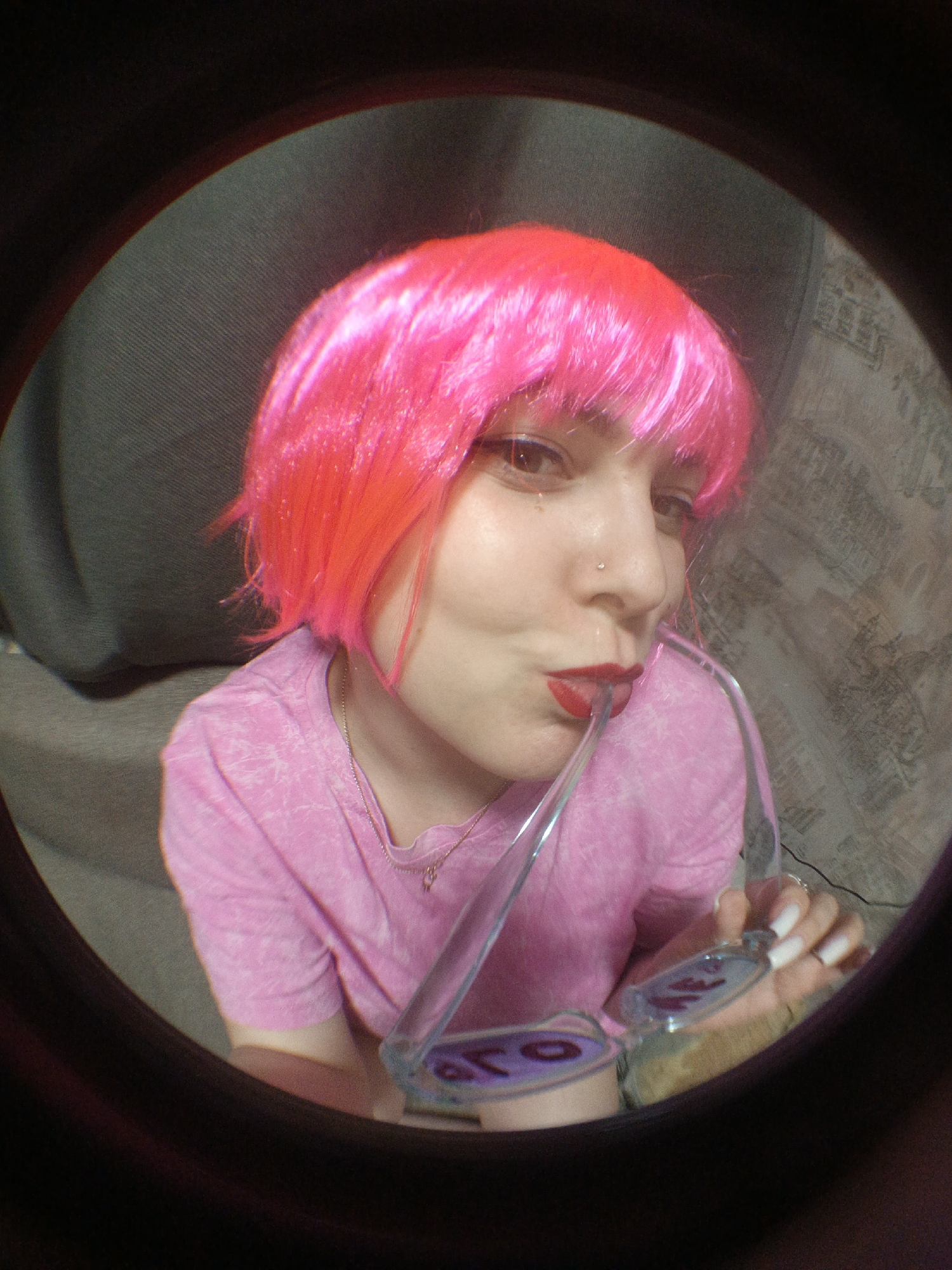 A person with light skin, a nose piercing, red lipstick, and a short, bright pink wig is depicted in a fisheye lens photograph. They are wearing a pink t-shirt and holding a pair of transparent sunglasses, placing the arm of the glasses in their mouth. The background is indistinct, showing a grey sofa and a patterned wall. The exact location within the city of Марковский, Russia cannot be determined from the image.
