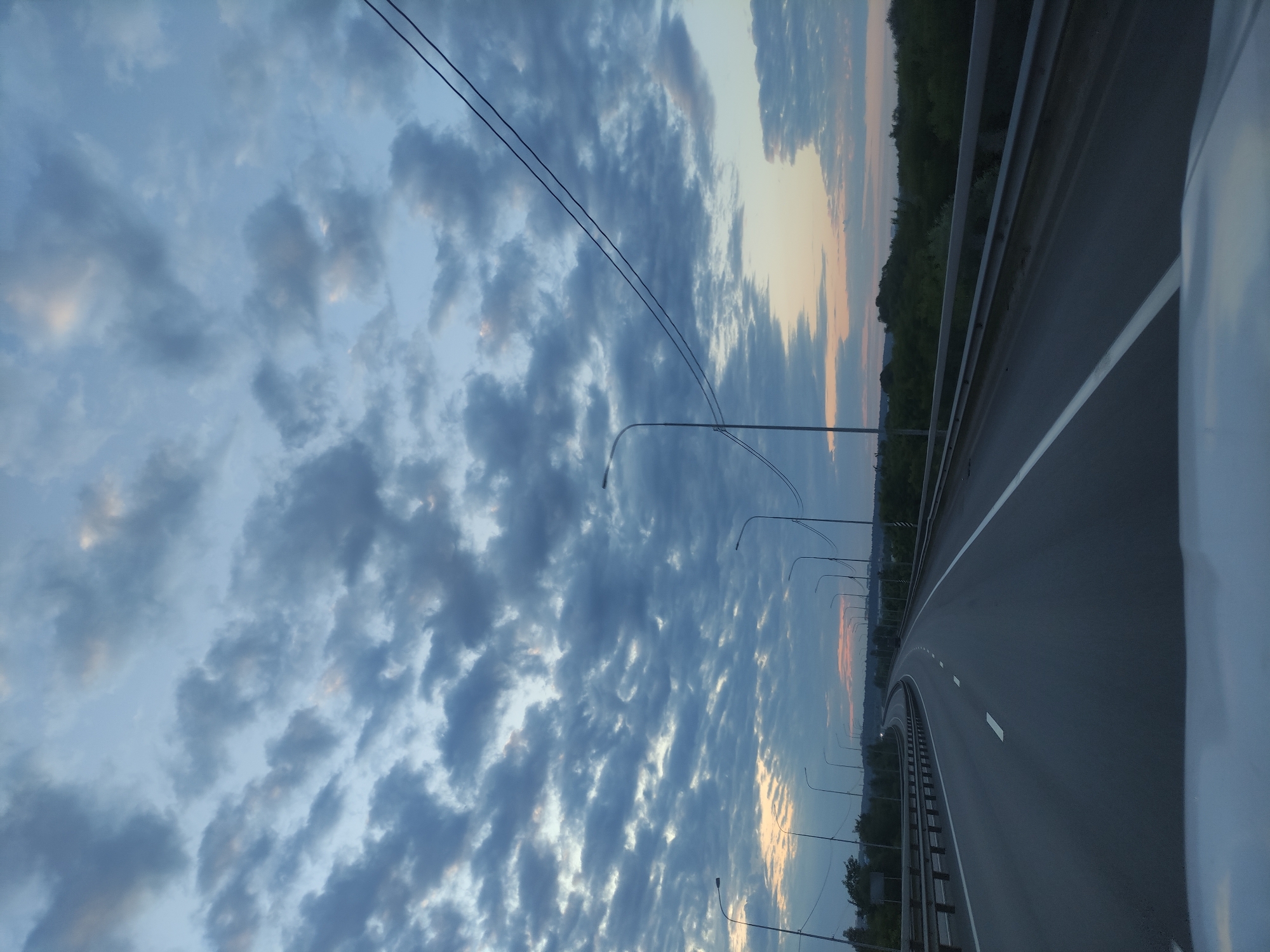 The image captures a wide-angle view of a highway under a vast, cloudy sky, likely taken from inside a vehicle during dawn or dusk. The sky occupies the majority of the frame, filled with numerous textured clouds ranging in color from deep blue-grey to lighter white and soft orange-pink, especially near the horizon. This color gradient suggests the sun is either rising or setting.
The asphalt highway runs across the lower and middle sections of the image, gently curving towards the right. It features clear white lane markings, including both solid and dashed lines. A metal guardrail stretches along the right side of the road. A line of dark green trees or foliage is visible in the distant background, bordering the horizon. A series of tall, slender streetlights or lamp posts line the road on the left, receding into the distance. Two thin, parallel overhead wires are visible against the sky in the upper left. A small, white portion of a vehicle's interior or body is visible along the far right edge of the frame. No people or visible text are present in the image.