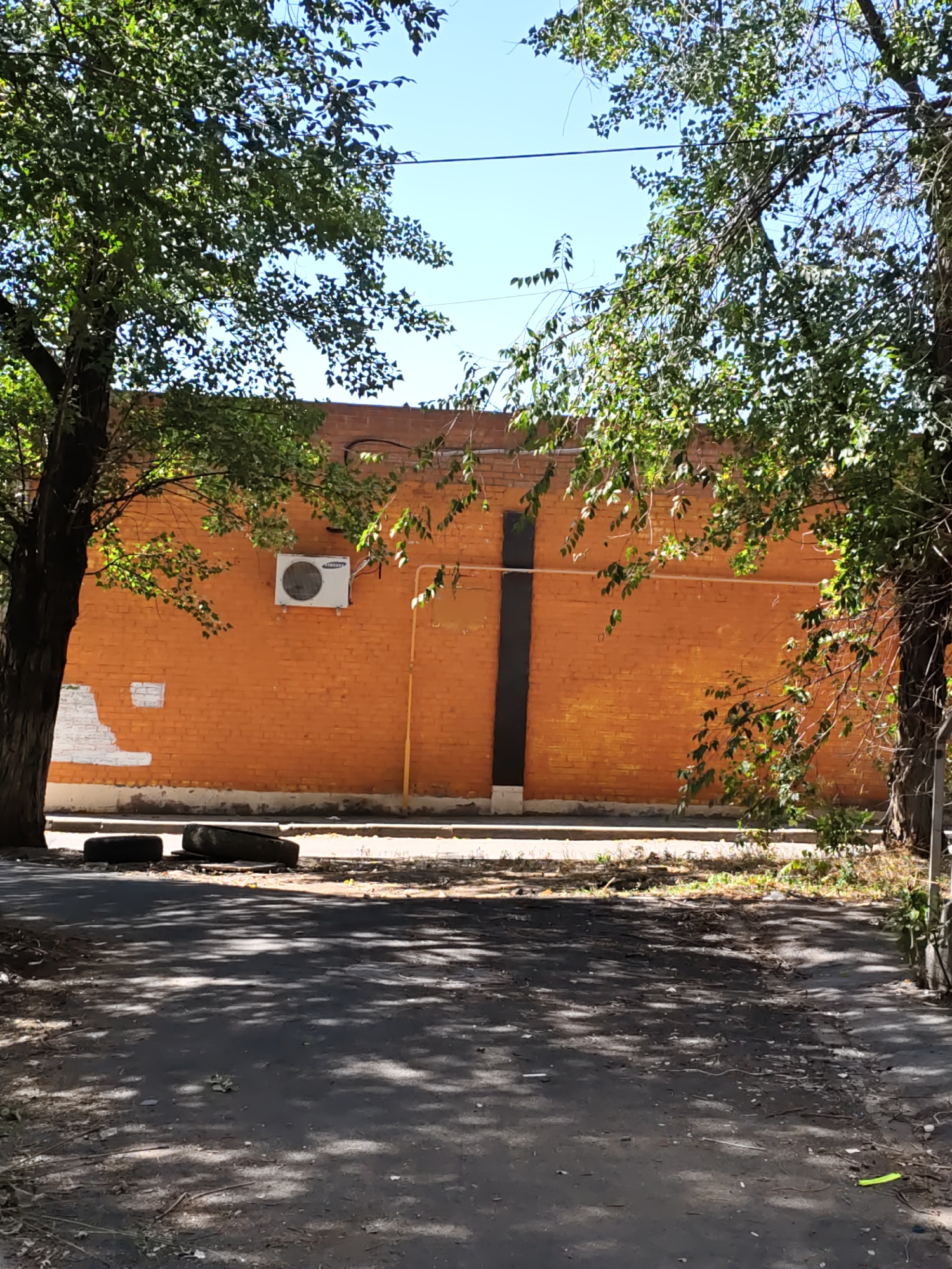 A sunny, outdoor scene in Rostov-on-Don, Russia, features a bright orange brick building wall partially obscured by the leafy branches of two large trees framing the view. An air conditioning unit is mounted on the wall to the left of a vertical black rectangular panel. Two worn tires are placed on the ground in front of the wall. The foreground shows a paved area with dappled sunlight and shadows, with some debris scattered on the surface. A yellow pipe runs vertically along the orange wall.
