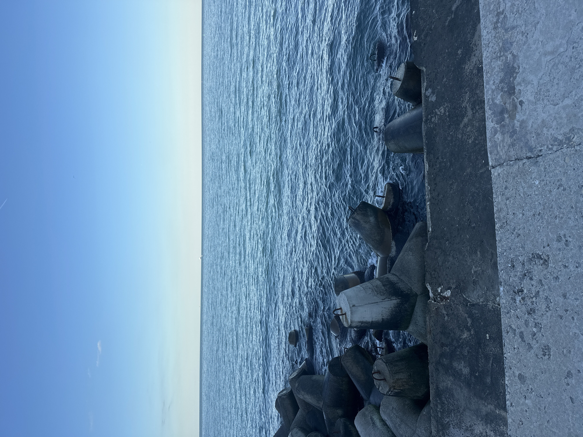 The image shows a concrete breakwater structure along the edge of the sea. The breakwater is composed of large, irregularly shaped concrete blocks, some of which have metal loops protruding from them. The sea is a deep blue, with small waves creating ripples on the surface. In the distance, the horizon is visible as a thin line where the sea meets the pale blue sky. There are no people or animals visible in the scene. The environment appears to be calm and overcast, with a muted color palette dominated by blues and grays.