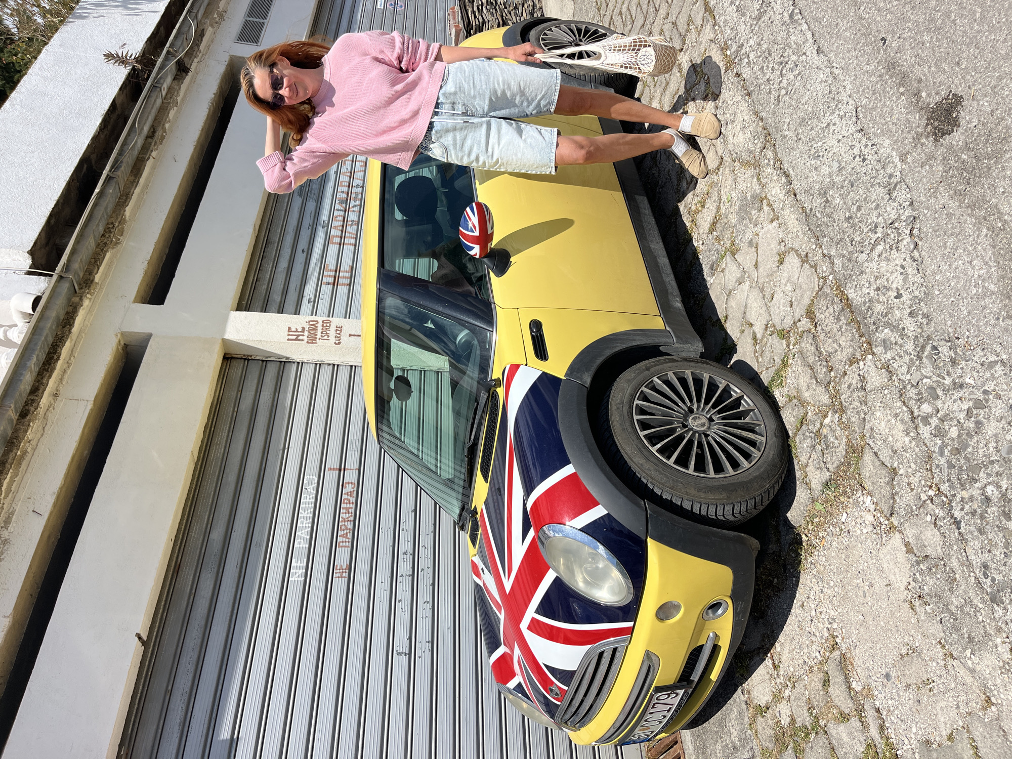 A fair-skinned person stands next to a yellow Mini Cooper car with a Union Jack design on its hood, posing in the city of Budva, Montenegro. The individual is clad in a pink sweater, light blue shorts, and neutral-colored shoes, and is wearing sunglasses. They are holding a white mesh bag. The car is parked in front of a building with closed roller shutters, and a sign with the inscription “NE PARKIRAJ ISPED GARAZE” is visible. The setting appears to be a sunny day on a paved street.