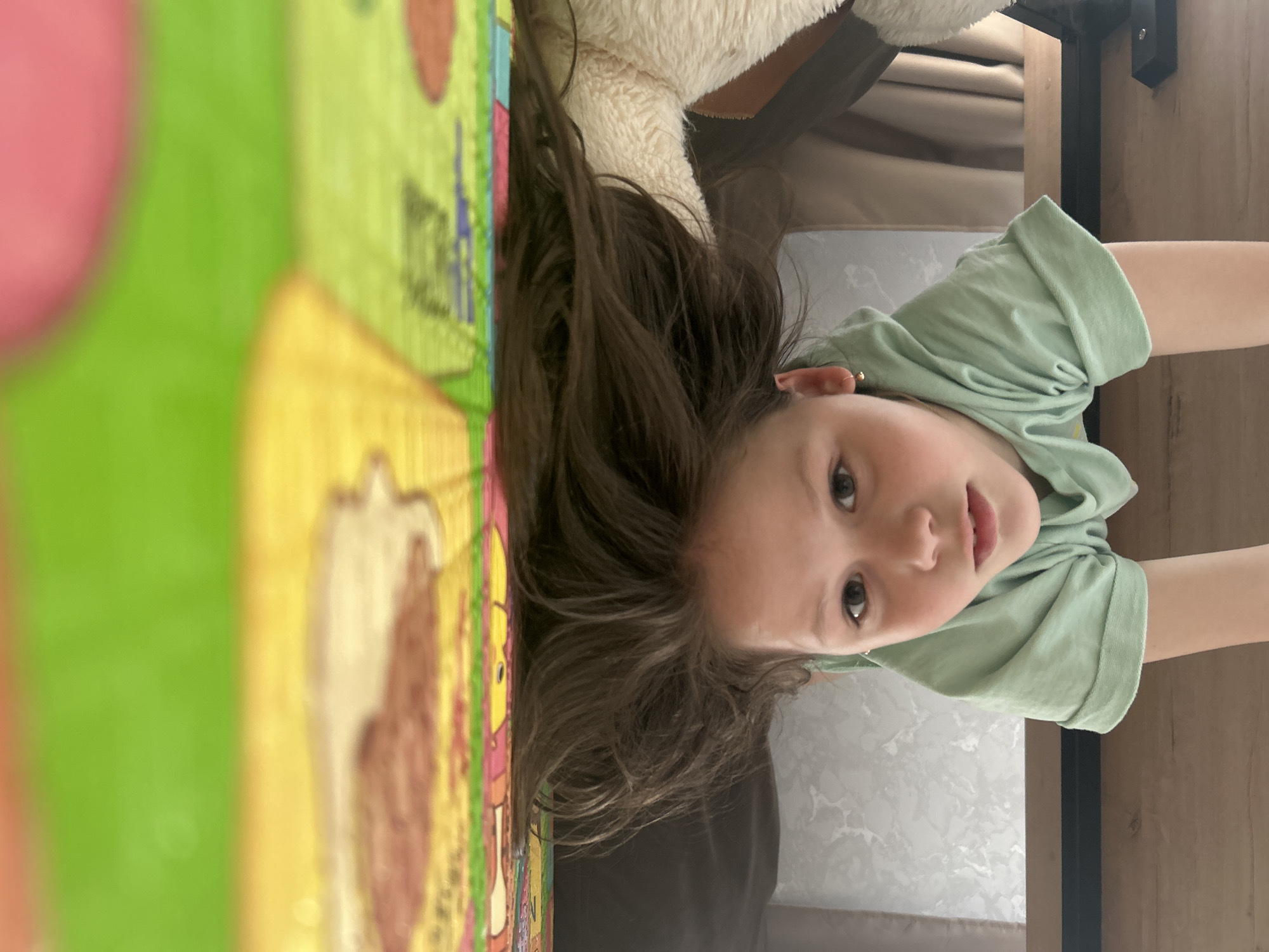 A young girl with long brown hair is looking at the camera, upside down. Her head is positioned over a colorful children's play mat with various images on it. She is wearing a light green T-shirt. A portion of a stuffed animal with white fur is visible above her head. In the background, there is a light-colored upholstered surface and a wooden surface. The setting appears to be indoors, possibly a home. The location, Chaikovsky, Russia, is not discernible from the image alone.